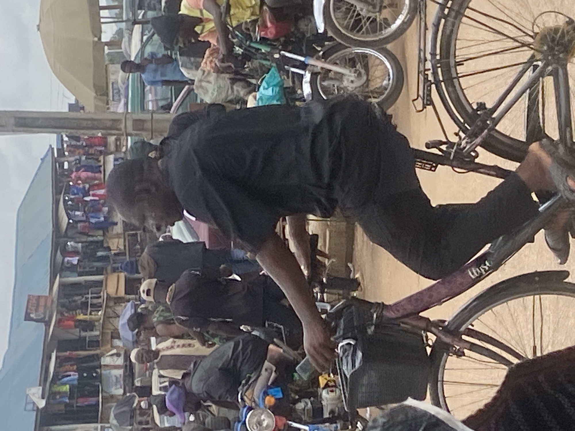 The image shows a street scene in Bukuru, Nigeria. In the foreground, a person is riding a bicycle. The person is wearing a black outfit. The bicycle has a basket in the front and a rack in the back. In the background, there are several motorcycles and a market area with stalls selling clothing. Other people are visible in the background, some appearing to be customers and vendors. The environment suggests a busy market day.