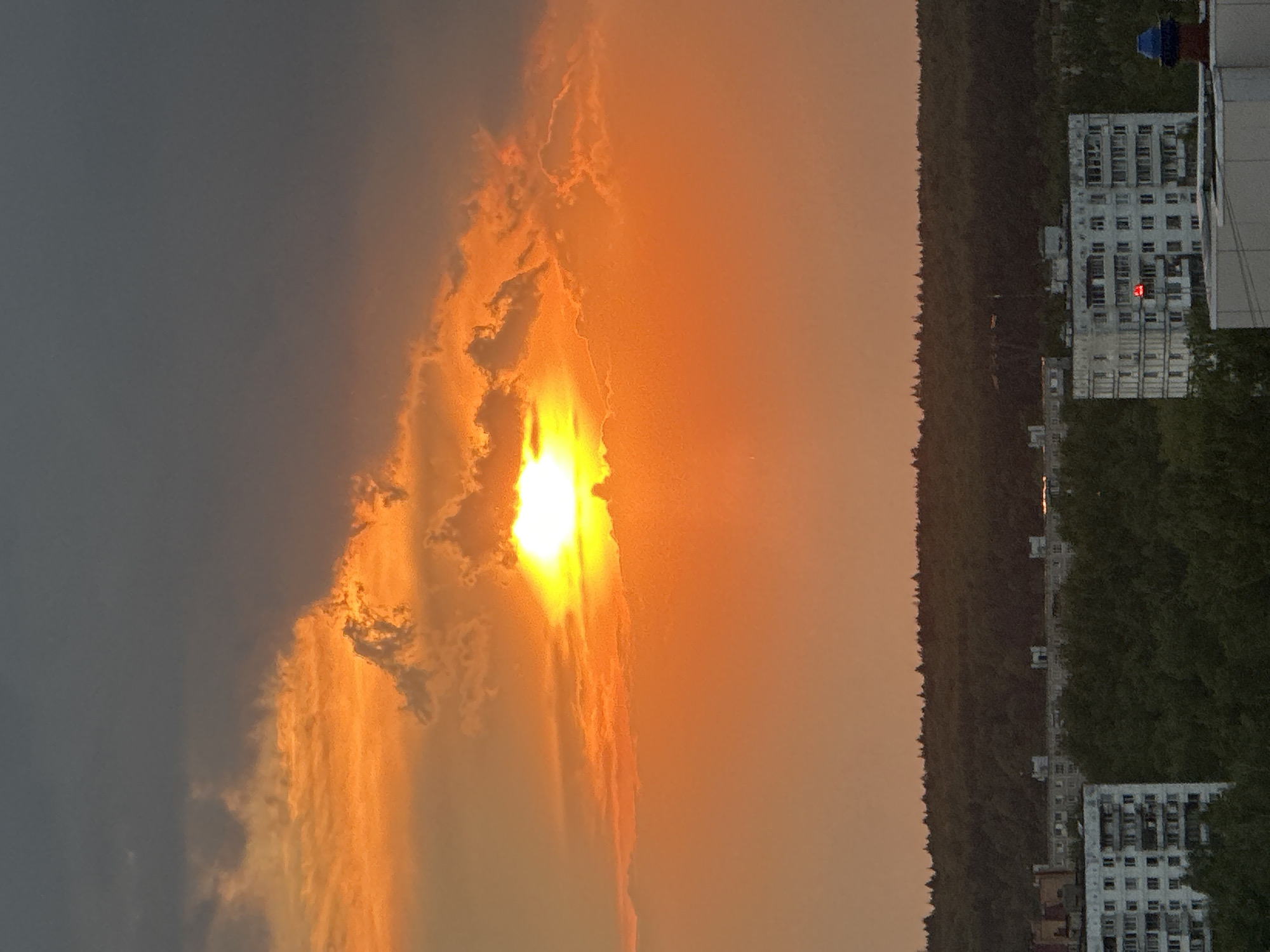 The image captures a sunset scene in Krasnogorsk, Russia. The sun is partially obscured by clouds, casting a bright orange and yellow glow across the sky. Adjacent to the skyline is a line of dense forest, beyond which are several residential buildings. Some of these buildings are high-rises with numerous windows, indicating apartment complexes. One particular high-rise has a red light visible on one of the upper floors. The foreground of the shot is primarily sky, transitioning from dark grey clouds on the left to the sunlit clouds on the right.