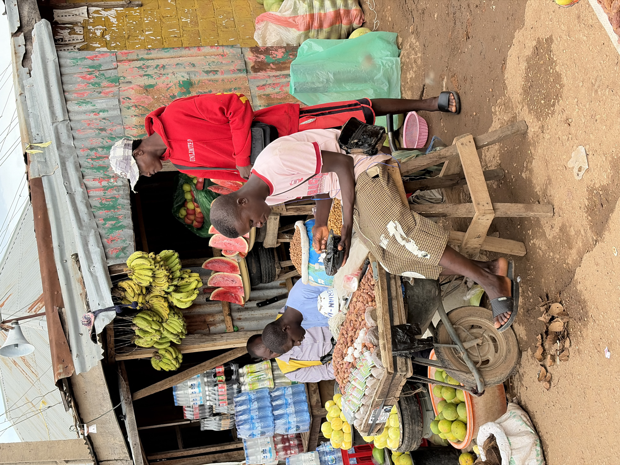The image depicts an outdoor market scene in Gura Top, Nigeria. In the foreground, a young man wearing a pink t-shirt and brown patterned shorts sits on a wooden stool, engaged with a tray of nuts. He is holding a black plastic bag and appears to be sorting or examining the nuts. To his right, another young man in a red hooded sweatshirt and red shorts stands, looking away from the camera. Behind them, a vendor in a striped shirt and yellow vest is visible behind a cart laden with various goods, including water bottles, fruits, and what appear to be nuts or seeds. The market stall features a corrugated metal roof and colorful, somewhat worn decorations. Various fruits, including bananas and watermelons, are displayed for sale. Bags of produce and scattered coconut shells are also present in the environment. The ground is dusty and uneven, suggesting a typical street market setting.