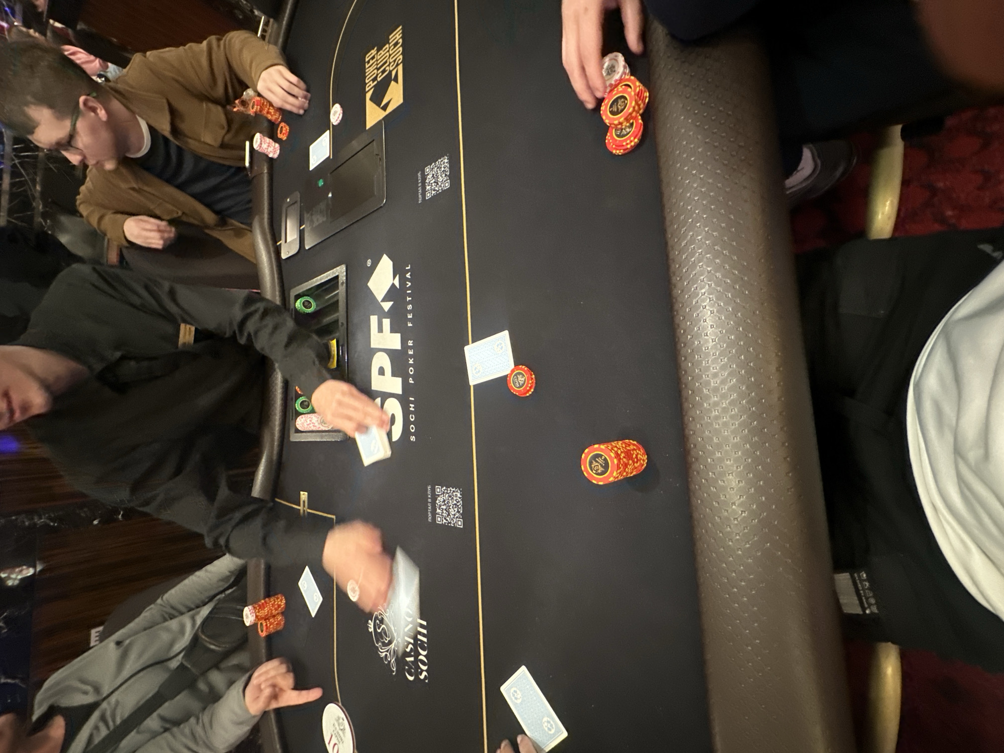 The image depicts a poker game in progress at a poker table, likely within the Casino Sochi in Estosadok, Russia, as indicated by branding on the table. Four individuals are partially visible around the table. Two men are seated on the right, one wearing a brown jacket and glasses, the other in a black shirt. Another person, wearing a gray jacket, is seated on the left. The fourth person, whose hands are active in the frame, appears to be a dealer or another player. The table is covered with a black felt surface, adorned with logos for "Casino Sochi Poker Festival" and "Poker Club Sochi." Various stacks of poker chips in red and yellow hues are distributed across the table, along with playing cards. One card is partially obscured by a hand. A QR code is also visible on the table. The overall setting suggests a formal or semi-formal gambling environment.