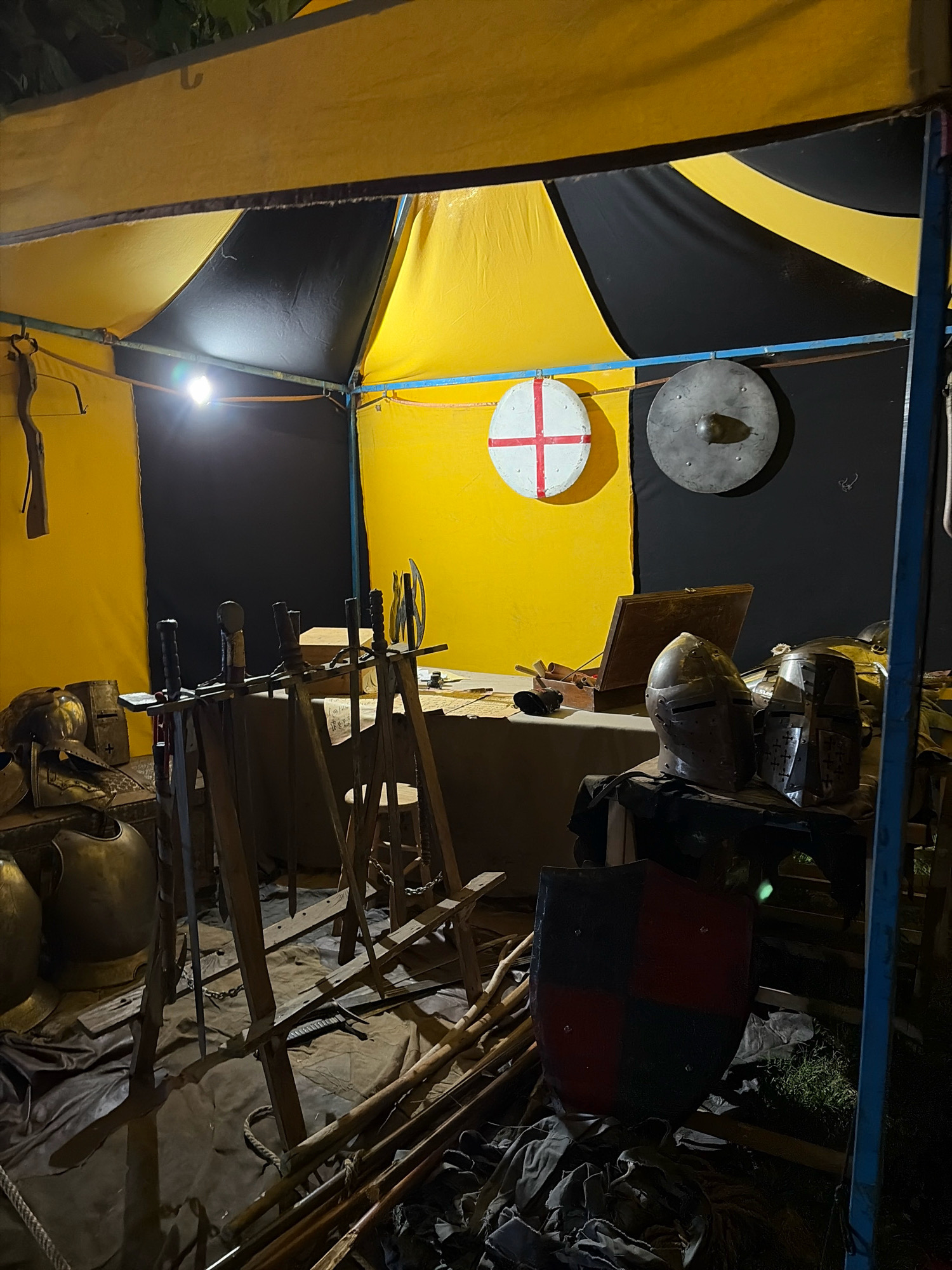 The image depicts a stall at a medieval-themed event, likely at the annual Roman and Medieval Market in Salou, Spain, held in the city center. The stall is set up under a yellow and black tent. Displayed are various medieval-themed items including suits of armor, helmets, swords, and shields. Two round shields are mounted on the back wall, one with a red cross on a white background, and another plain metal one. In the foreground, a red and black checkered shield rests on the ground. A variety of swords and poles are arranged in a rack. The lighting suggests it is nighttime or a dimly lit indoor setting. There are no people visible in the frame.