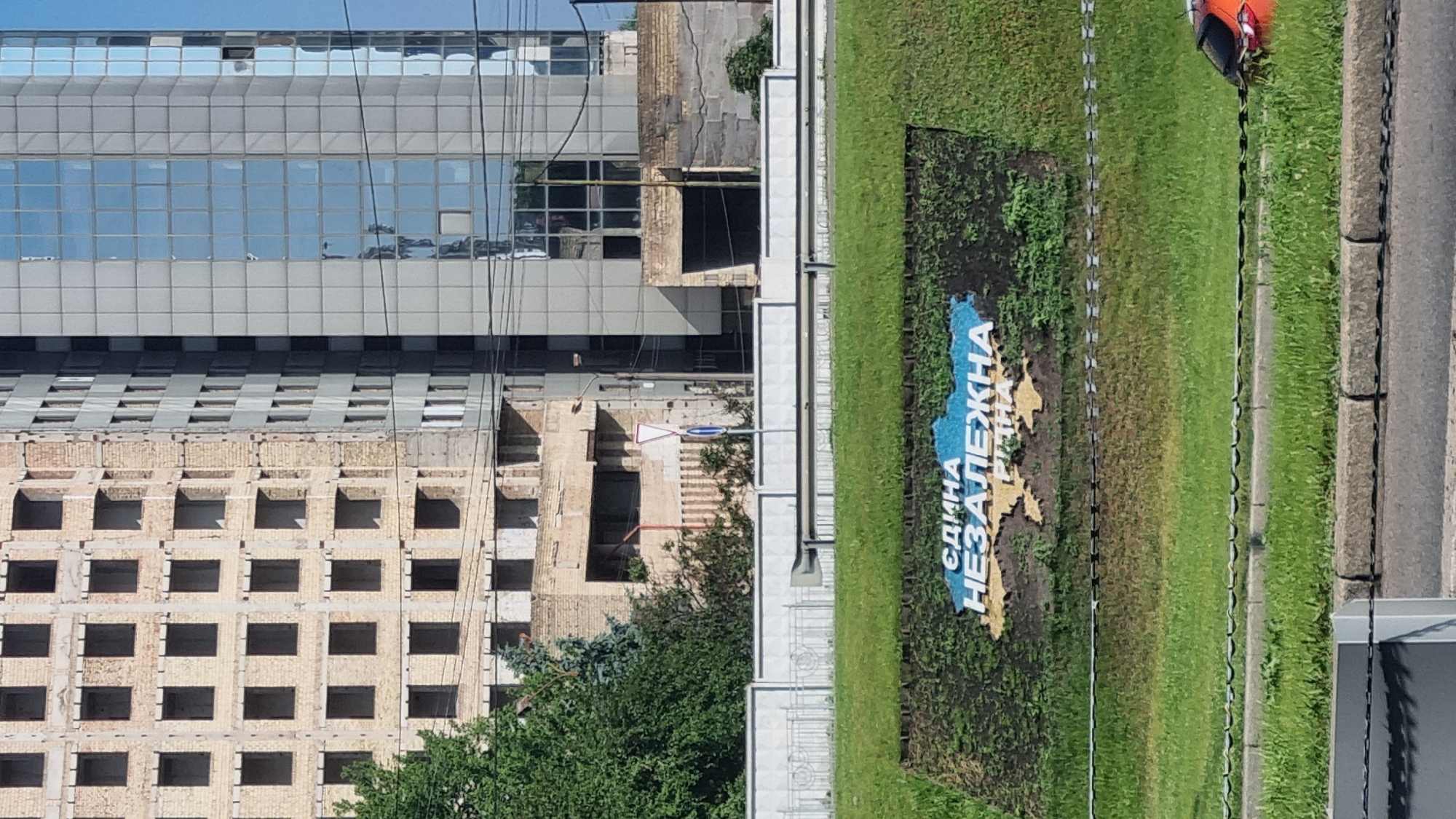 The image captures a scene in Kyiv, Ukraine, featuring a partial view of an unfinished building with exposed brickwork and numerous square openings that would have been windows. Adjacent to it is a modern glass-facade building. In the foreground, a grassy embankment displays a large mosaic depicting the outline of Ukraine, colored blue and yellow, with the text "ЄДИНА НЕЗАЛЕЖНА" (United Independent) written in large letters. The mosaic is set against a dark, textured background. A white fence runs horizontally across the middle ground, separating the embankment from a road or pathway. The overall environment suggests an urban setting, possibly with ongoing construction or development, and a clear expression of national pride through the mosaic.