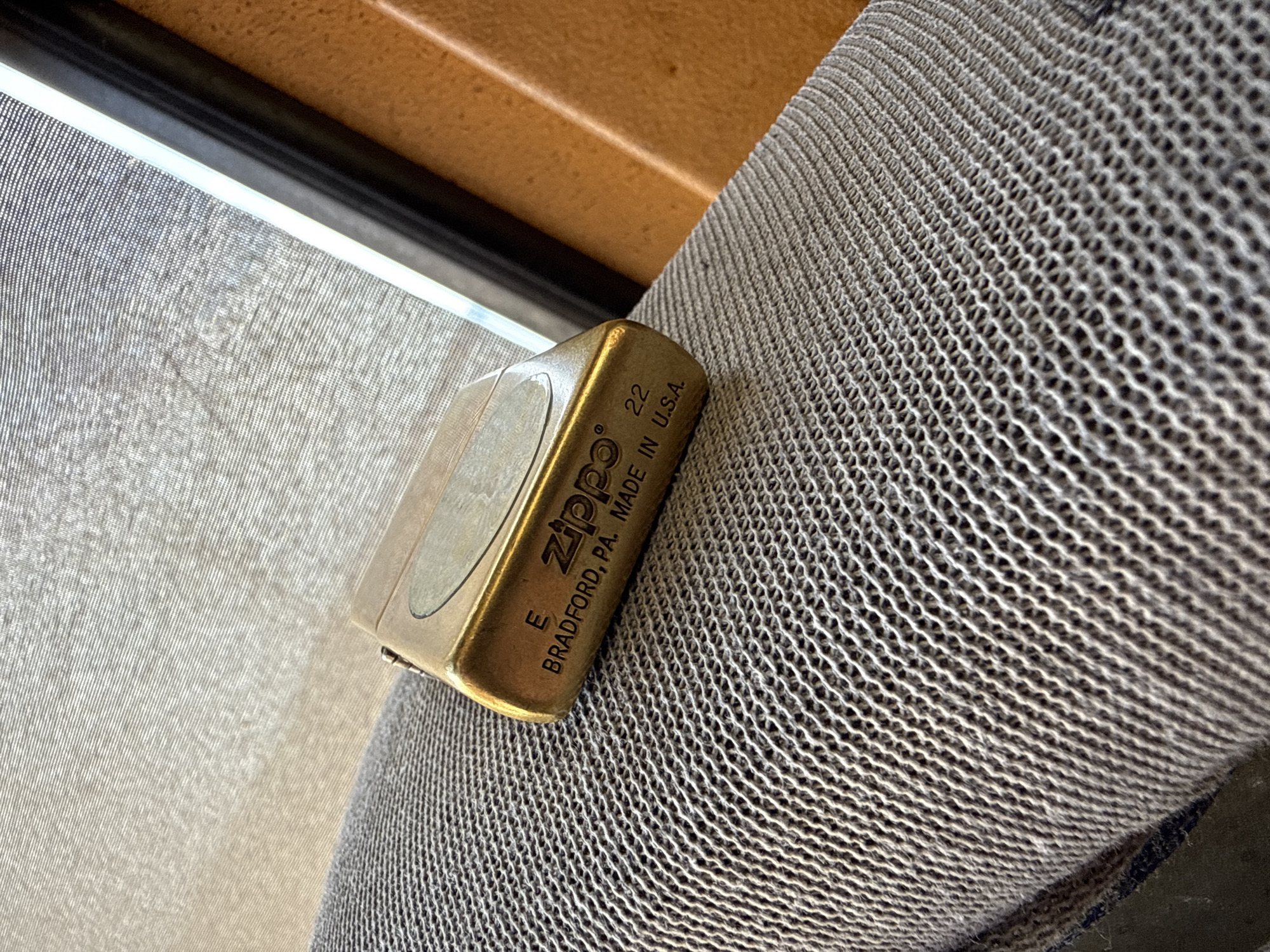 A Zippo lighter rests on a textured, grey surface, possibly a fabric or mesh material. The lighter is made of brass and has some text engraved on its surface. The text reads "ZIPPO 22 BRADFORD, PA. MADE IN U.S.A." and also "E". The lighting suggests it is daytime, with light filtering from an unseen window or opening. The background is blurred, showing a tan colored surface, and a dark, thin line that could be part of a frame or edge. The overall impression is one of a still life or object study, with the Zippo lighter as the central subject. The context of Mikołajki, Poland, suggests this might be a personal item within a home or outdoor setting in that location. There are no people or specific events depicted.