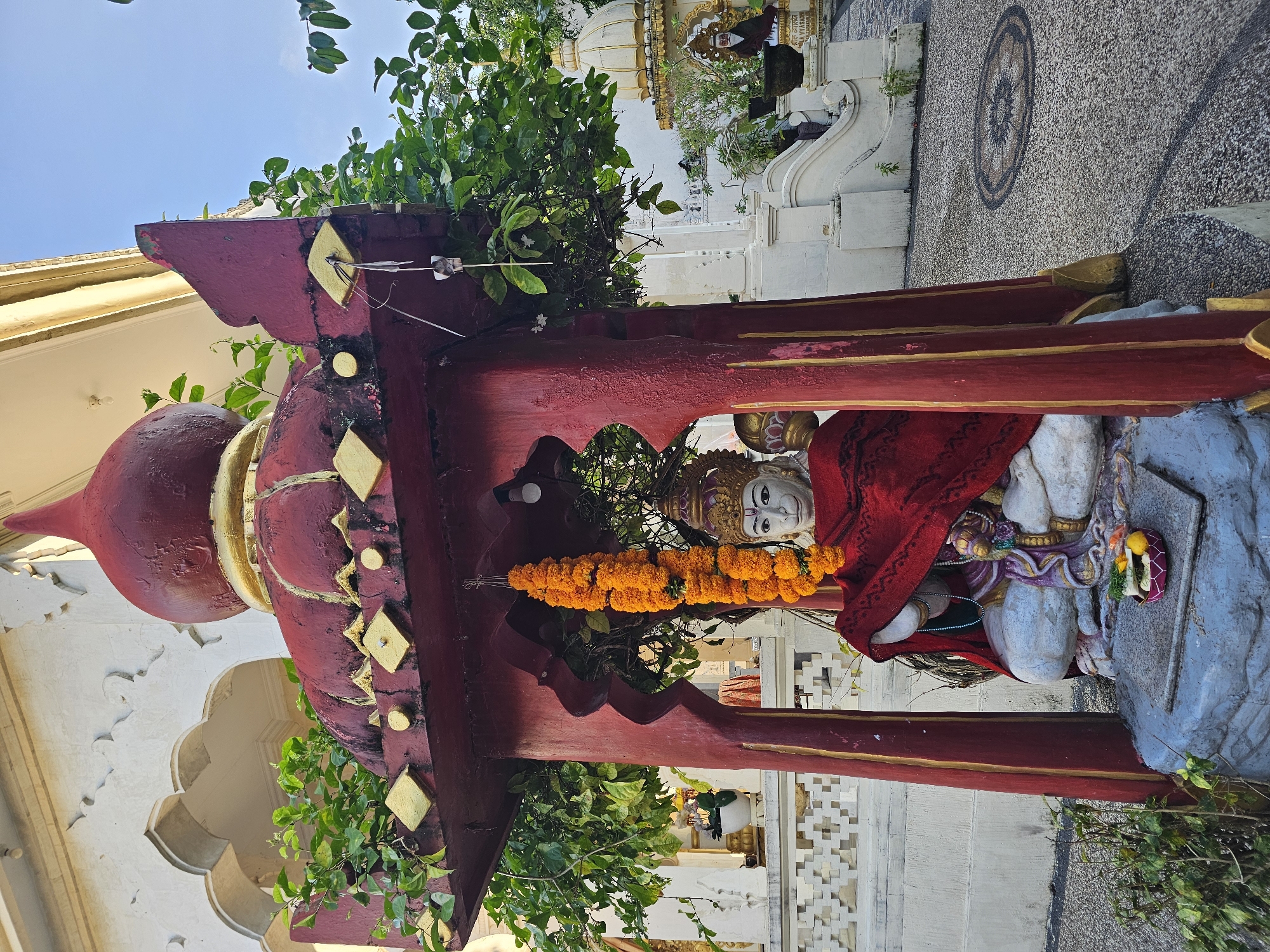 The image captures a statue of a deity at Pura Maospahit, a Hindu temple in Denpasar, Indonesia. The statue is adorned with a garland of orange flowers and draped in a red cloth. It sits beneath a red and gold painted structure that appears to be part of the temple architecture. Green foliage surrounds the statue and structure. The background reveals details of the temple complex, including white buildings and decorative elements. The ground is visible, showing a mosaic pattern and stone pavement. The overall setting suggests a sacred space within a traditional Balinese temple.