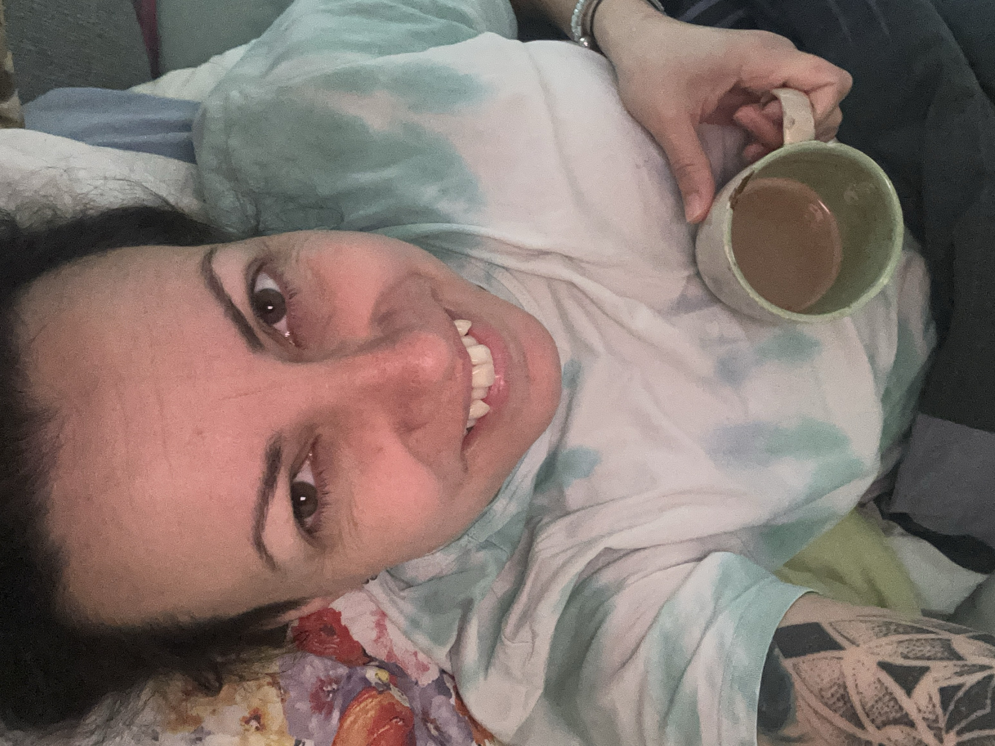 A fair-skinned woman with dark hair and brown eyes is lying down and smiling at the camera. She is wearing a tie-dye shirt and has a tattoo on her left forearm. Her right hand holds a light green ceramic mug filled with a brown liquid, likely coffee or tea. The setting appears to be indoors, possibly a bed or couch, as indicated by the presence of soft bedding and cushions. The background is out of focus, making it difficult to discern specific details about the environment. There is no information to indicate the location is in Sofia, Bulgaria.