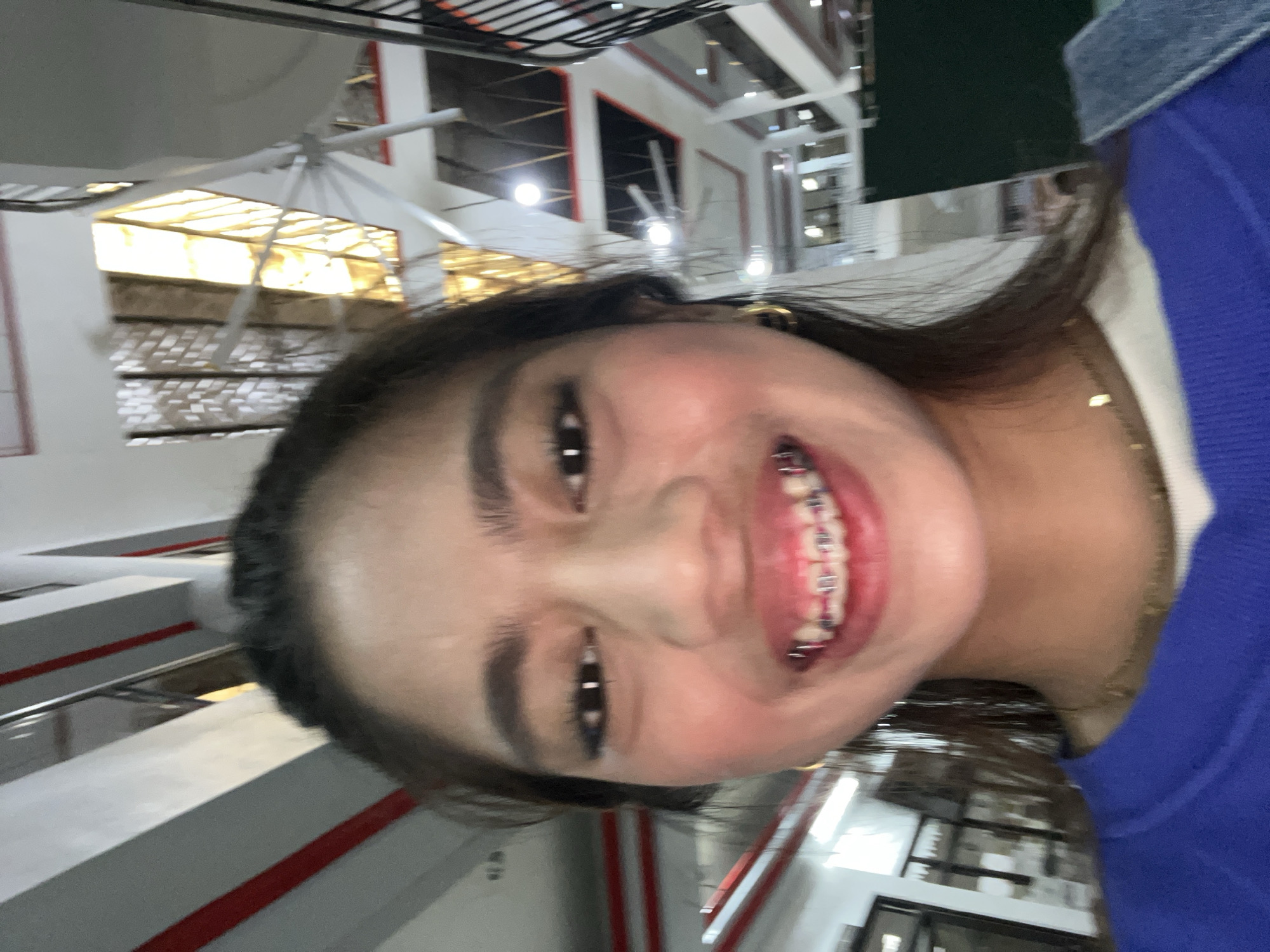 A young woman with braces smiles at the camera, her face filling most of the frame. She is wearing a blue sweatshirt and a gold necklace. The background is a blurry interior of a building with multiple levels and industrial-looking elements like exposed pipes and ceiling fans. The building appears to be a public space, possibly a mall or a government building, with concrete structures and large windows or openings that let in natural light. The image is taken from a low angle, looking up at the woman. There is no identifiable signage or landmarks that specifically place the location in Panabo City, Philippines.
