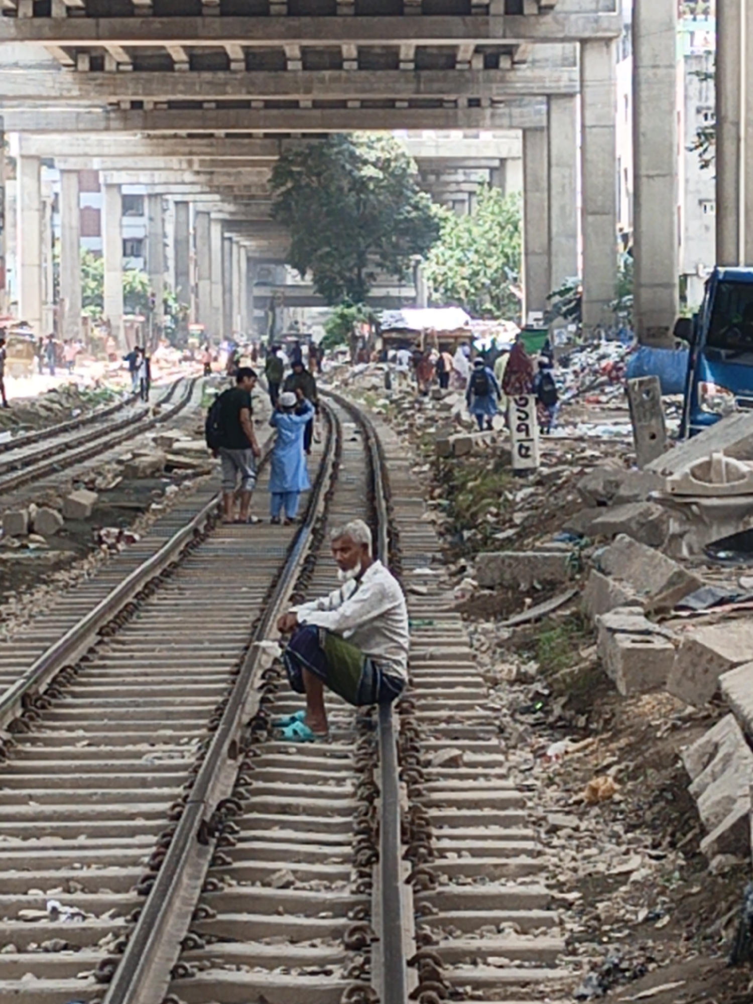 The image shows a scene in Dhaka, Bangladesh, featuring a man sitting on a railway track. The railway lines run through a cluttered urban landscape, with people walking along and across the tracks. Overhead, a concrete flyover structure dominates the background. Rubble, trash, and debris are scattered along the sides of the tracks and the surrounding areas. Several individuals can be seen walking or standing on the tracks, while others are visible in the background. The environment appears congested and impoverished.