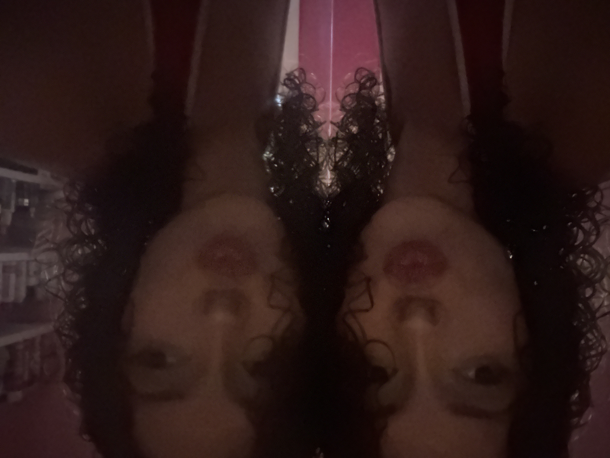 The image displays a symmetrical reflection of a young woman with curly dark hair. Her face is partially obscured, but her lips are parted and painted red. She appears to be wearing a red top. The background is dark and somewhat blurry, with hints of shelves and bottles suggesting an indoor setting, possibly a bathroom or a retail space. There is no discernible information indicating a specific location within Milan, Italy, in the provided image.