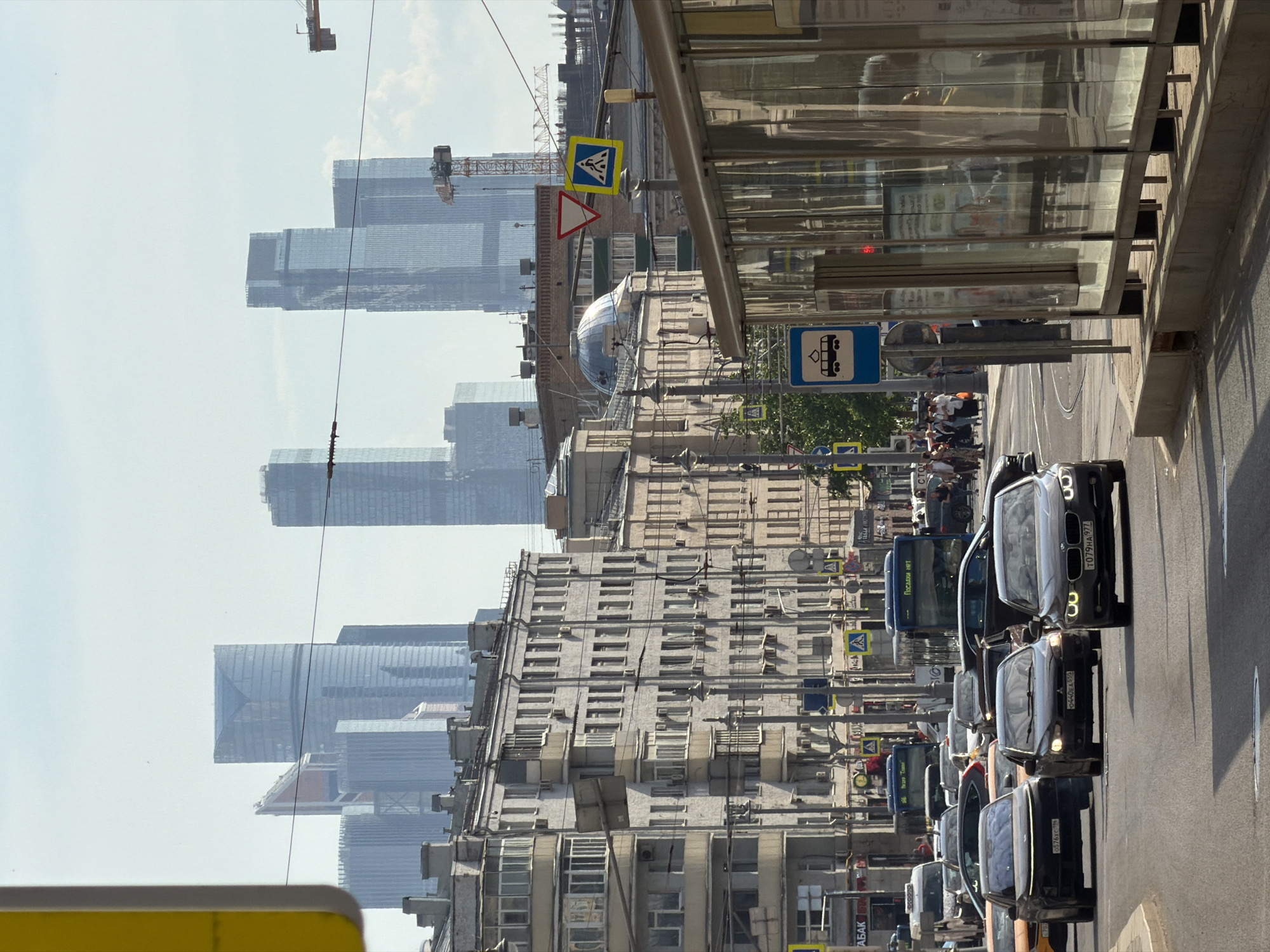 The image shows a street in Moscow, Russia, with modern high-rise buildings in the background. The street is lined with older buildings, pedestrian crossings, and bus stops. A line of cars and buses fills the street, moving slowly in traffic. People can be seen waiting at a bus stop. The sky is hazy, and there are construction cranes visible among the skyscrapers.