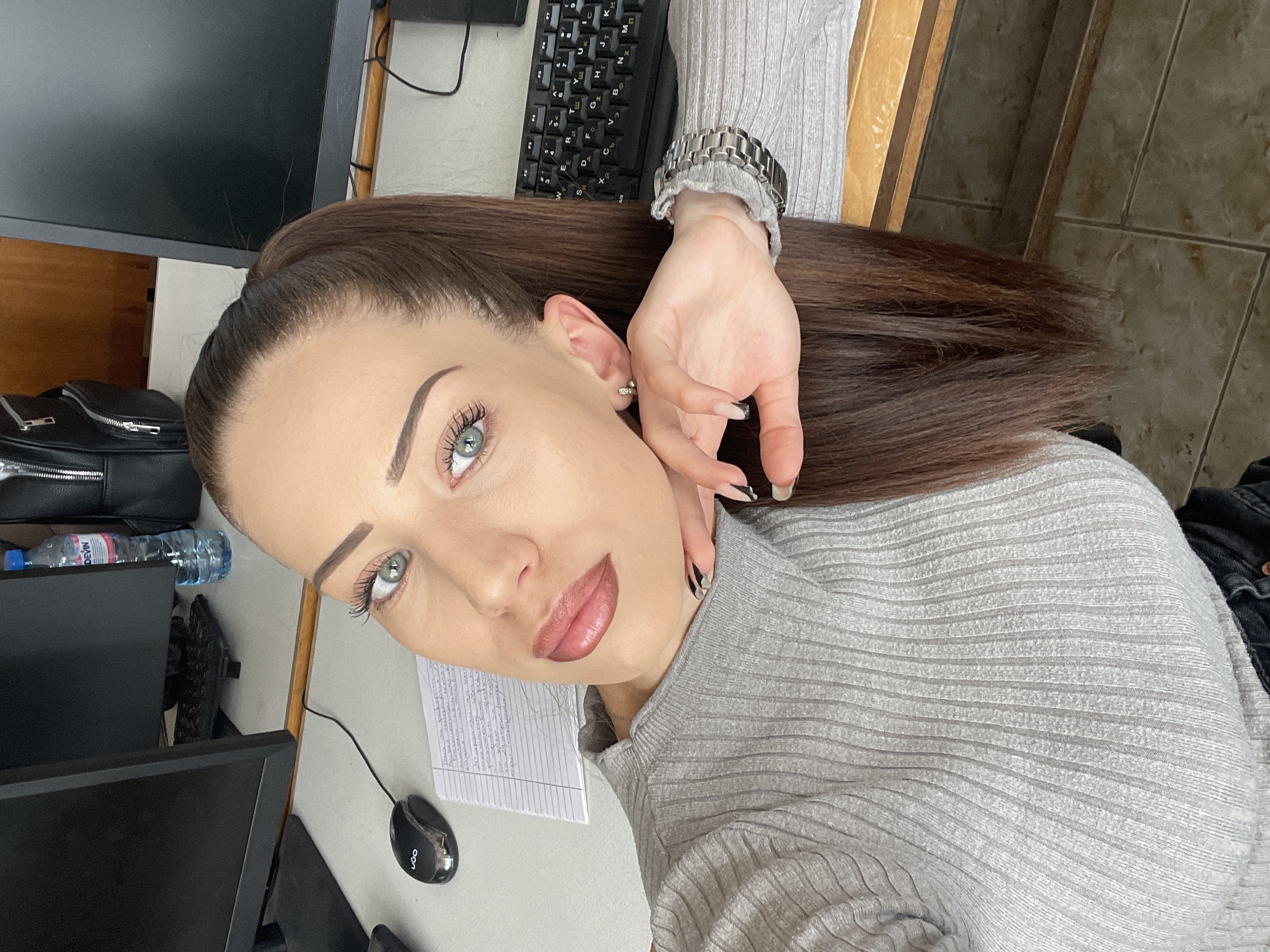 A light-skinned woman with brown hair pulled back in a ponytail is posing for a selfie. She has light green eyes, defined eyebrows, and full lips. Her left hand is touching her face. She wears a silver watch and a gray ribbed top. The background includes a desk with a computer monitor, keyboard, mouse, and a notebook with writing on it. A bottle of water and a black bag are also visible. The location is not discernible from the provided image.