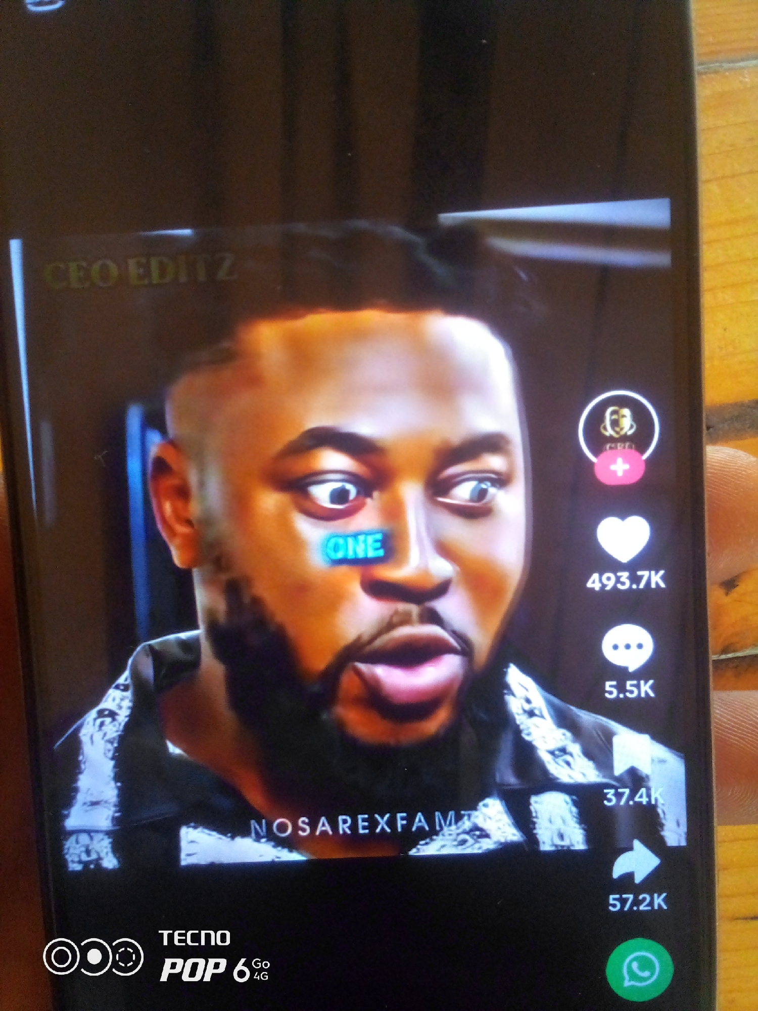 This is a close-up, vertical shot of a smartphone screen displaying a video. The video features a man with dark skin, a buzz cut, and a beard. He is looking directly at the camera with wide, surprised eyes. A bright blue rectangle with the word "ONE" in white letters is superimposed over his cheekbone.
On the right side of the screen, there are several social media interaction icons. At the top, there is a circular profile icon with a figure inside, and a pink plus sign below it, likely indicating a follow button. Below this is a white heart icon with the number "493.7K" beneath it, suggesting likes. Further down, there is a speech bubble icon with "5.5K" next to it, likely representing comments. Below that, a ribbon icon with "37.4K" is visible, possibly indicating shares or saves. Finally, at the bottom right, a green WhatsApp icon is displayed with the number "57.2K" beneath it.
At the top left of the screen, the text "CEO EDITZ" is visible in a yellow font. Near the bottom center, the text "NOSAREXFAM" is displayed in white, with some glitched or distorted elements overlaying it. In the bottom left corner, the "TECNO POP 6 Go 4G" logo is visible.
The background of the video appears to be a dark, possibly indoor setting. The smartphone itself is being held, with a portion of a finger visible on the right side of the image. The overall impression is of a social media video with engagement metrics and branding elements. The user's expression suggests an element of surprise or shock within the video content. The presence of "Howayi, Nigeria" in the context suggests this video might be related to content popular in that region.