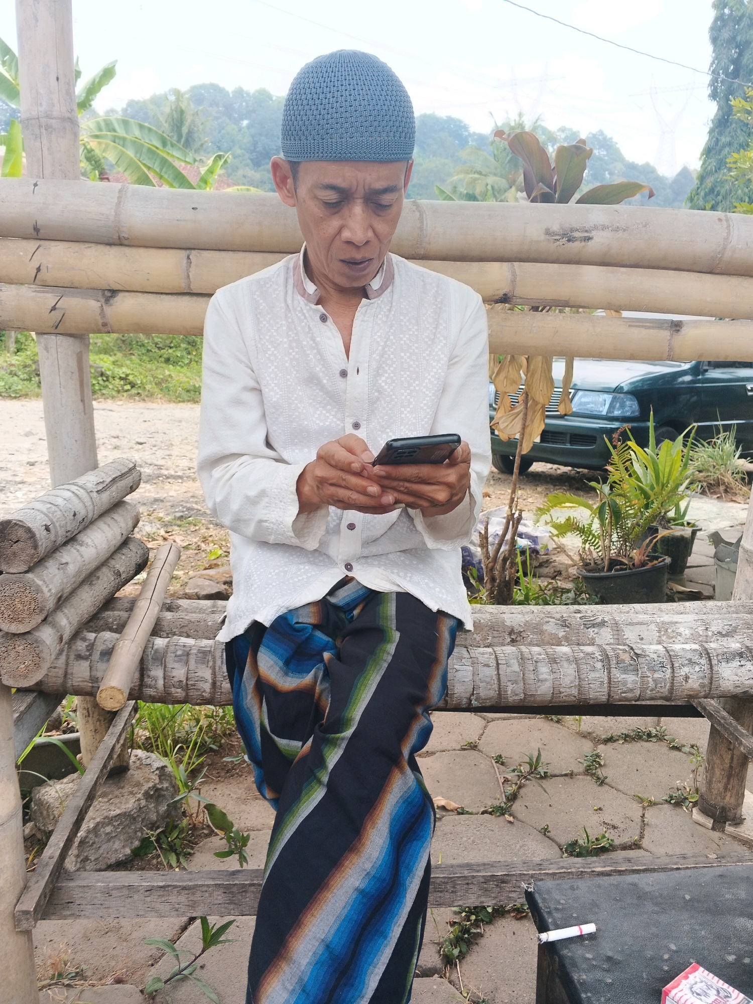The image shows a man wearing a white shirt and a blue cap, sitting on a bamboo structure and looking at his phone. He is also wearing a sarong with blue, black, and white stripes. The background shows a natural environment with trees and a car. The scene appears to be in Ungaran, Indonesia, based on the cultural attire and the surrounding vegetation.