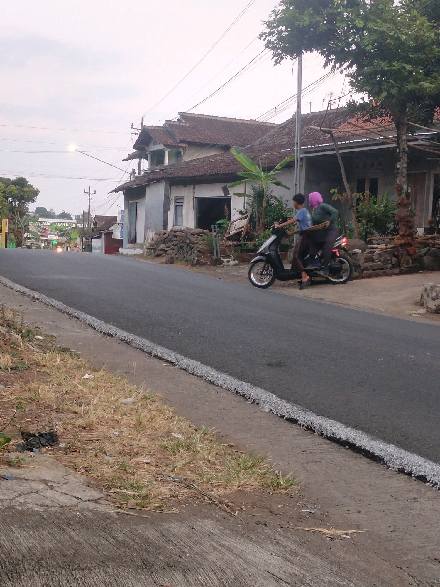 A low-angle, medium shot captures a rural road scene in Pujut, Indonesia, with a clear sky overhead. The asphalt road occupies the foreground, bordered by dry, sparse grass and concrete on the left. In the mid-ground, a black scooter carries two individuals: a male wearing a blue t-shirt and dark pants, and a female in a green jacket and pink headscarf, holding a flat, dark object. They are positioned on the right side of the road, near a building with a tiled roof and stone wall. A sign on the left reads "SELAMAT JALAN DESA PUJUT," indicating the location. The background reveals more houses with tiled roofs and dense foliage, suggesting a village setting. A streetlamp illuminates the scene from the distance.