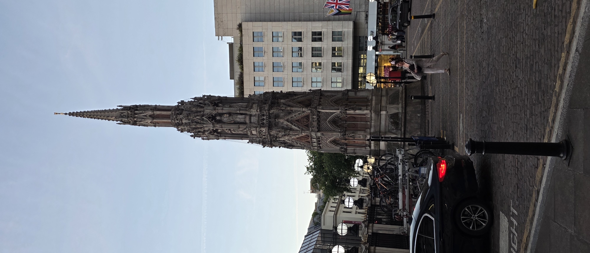 This image captures a view of the Albert Memorial in Kensington Gardens, London, United Kingdom. The memorial is a towering Gothic structure with intricate carvings and sculptures. In the foreground, a black car is parked, its brake lights illuminated. To the left of the memorial, a row of decorative lampposts illuminates a walkway. Several bicycles are parked on racks near the memorial. In the background, a modern multi-story building stands, its facade adorned with numerous windows. The sky is a pale blue with faint contrails. A woman in light-colored clothing is walking across the cobblestone street, carrying a bag.