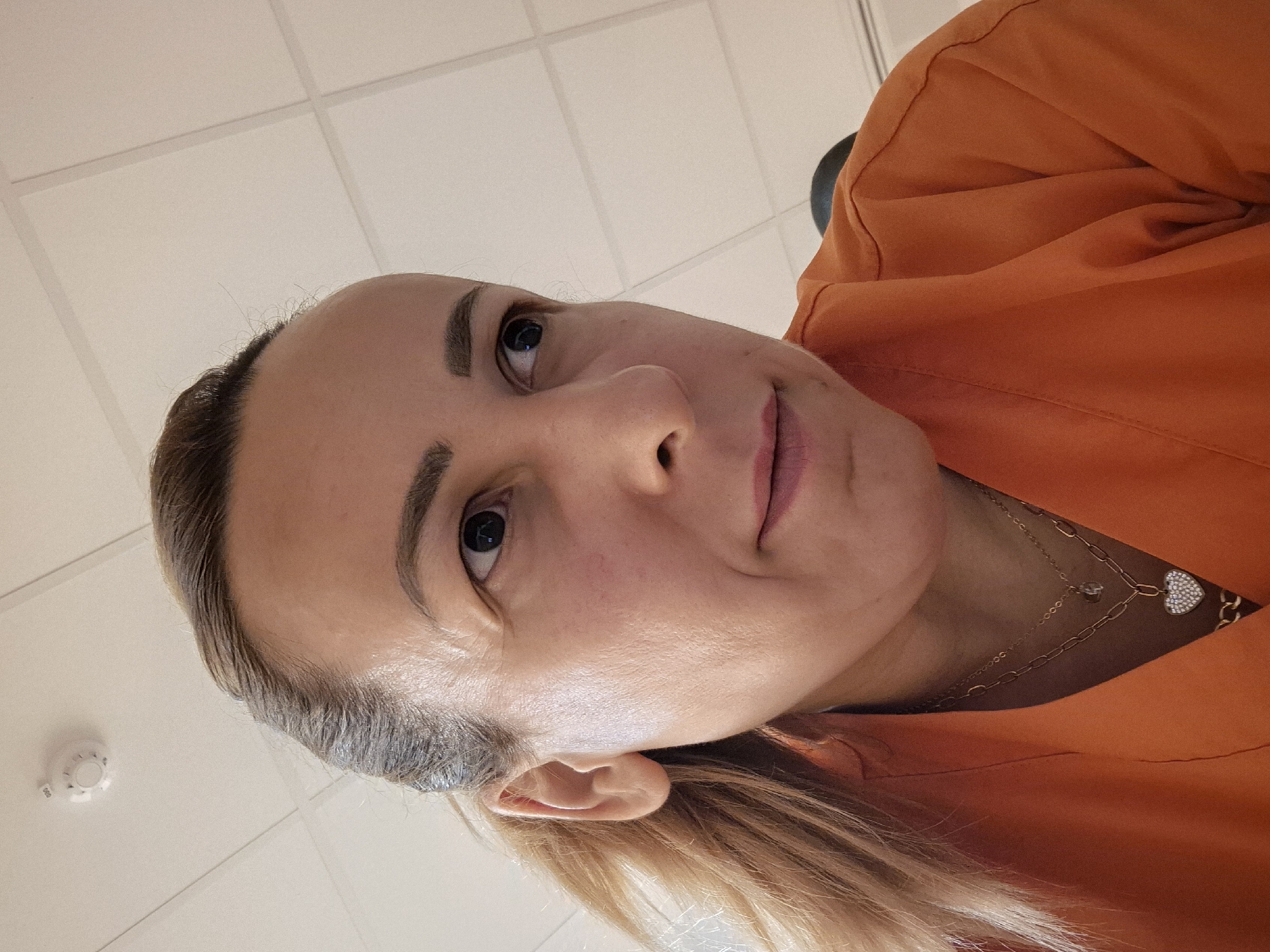 A close-up, low-angle, selfie-style shot shows a light-skinned woman with blonde hair pulled back. She is wearing an orange scrub top and a necklace with a heart-shaped pendant. She is looking to her right with her mouth slightly open. The background is a white tiled ceiling with a smoke detector visible in the top left corner. The lighting is bright and even.
