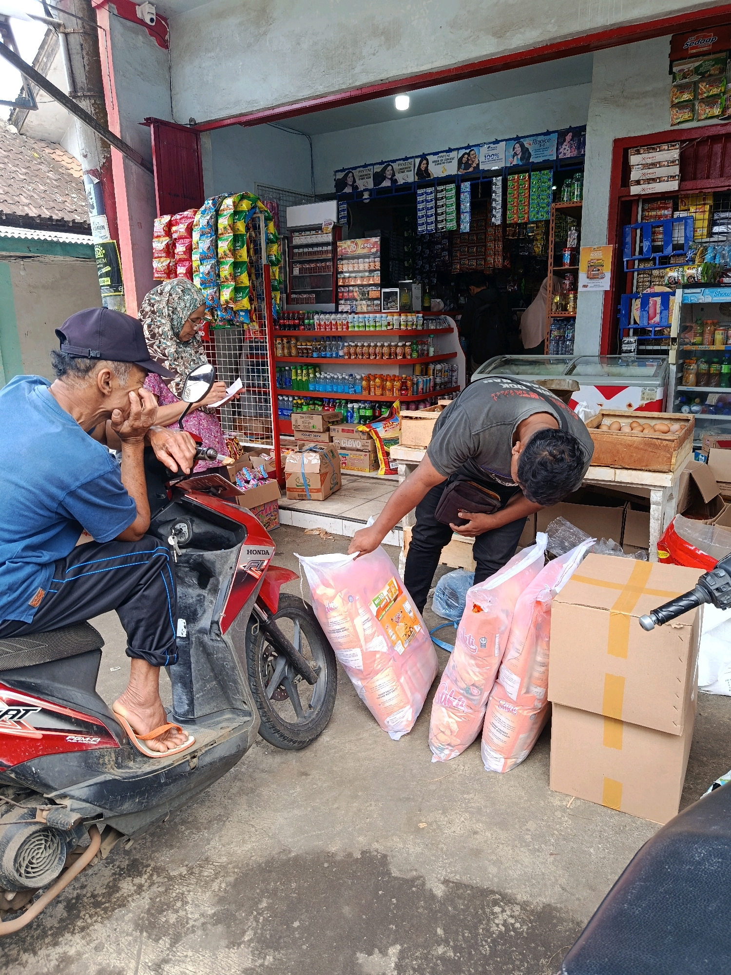 A man wearing a blue t-shirt and black pants sits on a red and black scooter in front of a small store. He is wearing a black cap and has his left hand on his face. Next to him, a woman in a floral print top and headscarf holds a piece of paper and appears to be talking to him. A third person, a man in a grey t-shirt and black pants, is bending down near the scooter, holding several large bags of what appear to be groceries. He is also holding a wallet in his left hand. The store behind them is stocked with various goods, including shelves of packaged food items, drinks, and what looks like a cooler. The ground is concrete, and there are several cardboard boxes and more bags of goods near the entrance of the store. The overall scene suggests a transaction or interaction at a local convenience store in Sidorenggo, Indonesia.