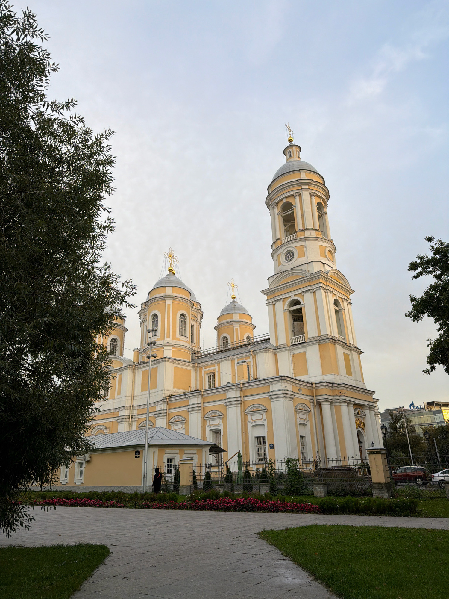 The image shows the Smolny Cathedral in Saint Petersburg, Russia, on a slightly overcast day. The cathedral is a prominent yellow and white building with several domes and a tall bell tower topped with a golden cross. The bell tower features arches and a clock face.
In the foreground, there is a paved pathway and a well-maintained lawn. A dark green tree with thin leaves partially obscures the left side of the cathedral. A small, low yellow building with a grey roof is situated in front of the main cathedral structure, separated by a metal fence. A person in dark clothing is visible standing near this smaller building.
Beyond the cathedral, modern glass-fronted buildings and trees can be seen, along with a red car and a white car parked on a street. The sky is a pale blue with some hazy clouds. The lighting suggests it is either early morning or late afternoon.