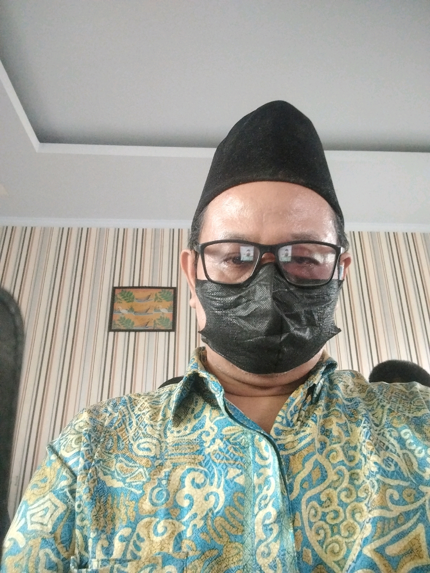 A selfie of a person wearing a black peci hat and a black face mask is taken from a medium close-up, chest-up angle. The person is also wearing a long-sleeved batik shirt with a pattern of blue and yellow swirls and geometric shapes. Behind the person, the wall is covered with vertical striped wallpaper in beige and light brown hues. A small framed artwork depicting boats and foliage hangs on the wall. The ceiling above is a plain white, recessed design. The image appears to have been taken indoors, possibly in a residential setting in Serang, Indonesia.