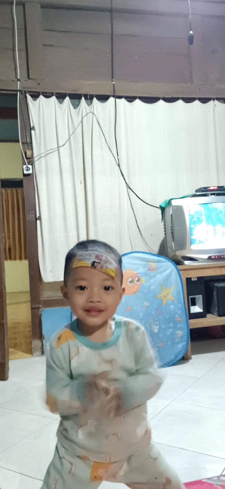 In Wonokerso, Indonesia, a young boy with a bandage on his forehead is captured in a candid indoor photograph. He is wearing light blue pajamas with a pale green collar and appears to be in motion, with his hands blurred in front of his chest. The background reveals a simple living space with white curtains hanging behind him. To the right, a vintage television set sits atop a wooden stand, accompanied by a blue toy bin decorated with fish and a starfish. The floor is tiled in a light color. The lighting suggests an indoor environment, possibly during the daytime, given the natural light filtering through the curtains.