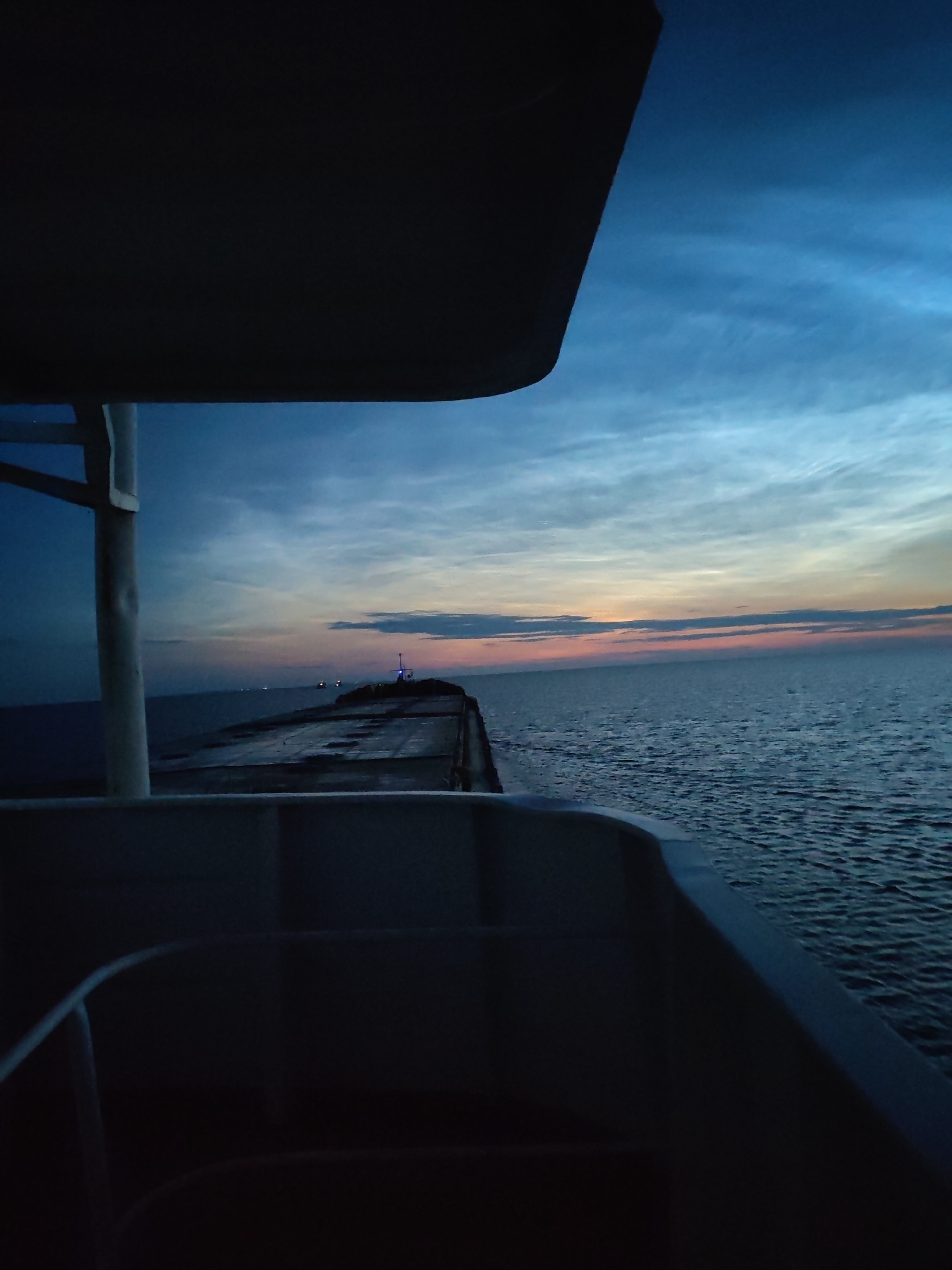 The image depicts a view from a vessel at dusk, looking out towards the sea. In the foreground, the dark silhouette of the vessel's interior is visible, with railing structures and seating areas. The mid-ground features a long, flat structure extending into the water, possibly a pier or breakwater, with a small navigational light or beacon visible at its end. The background is dominated by the expansive sea meeting the horizon, with the sky displaying a gradient of colors from deep blue at the top to soft oranges and pinks near the horizon, indicative of a sunset or twilight. Wispy clouds are scattered across the sky. The water is calm, with a gentle ripple. There are no people or animals clearly visible in the image. The scene suggests a serene, possibly remote location.