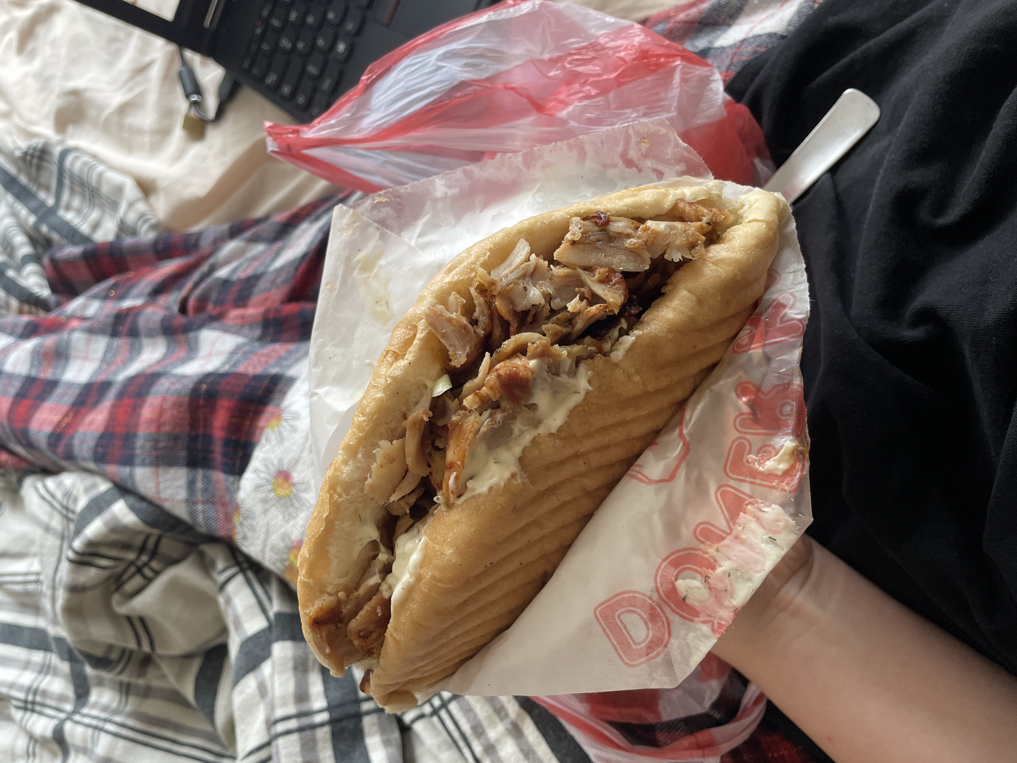 A person is holding a doner kebab sandwich in a paper wrapper. The sandwich is filled with meat and a white sauce. A red and white plastic bag is visible in the background, along with a laptop and other items resting on a bed or similar soft surface covered with checkered and solid black fabrics. The scene does not provide specific details to identify Zlatograd, Bulgaria.