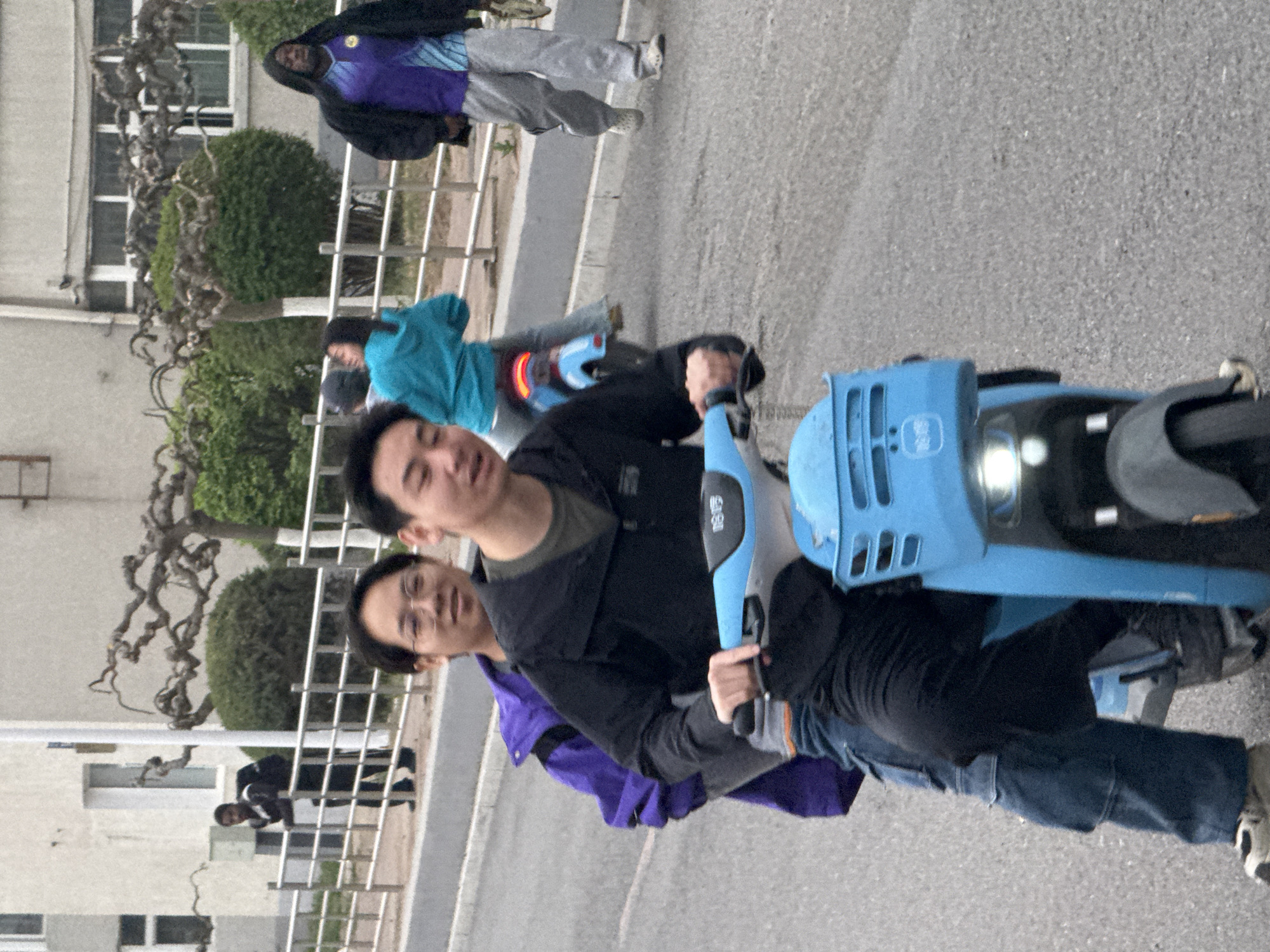 Two Asian males are riding a light-blue electric scooter on an asphalt road, near a sidewalk and greenery. The rider in front wears a black jacket and the passenger behind wears a purple jacket. Other people are nearby, walking or riding similar scooters. The location appears to be Zhongtun, China, as indicated by signage or markings on the scooter. Buildings and trees are visible in the background, suggesting an urban setting.