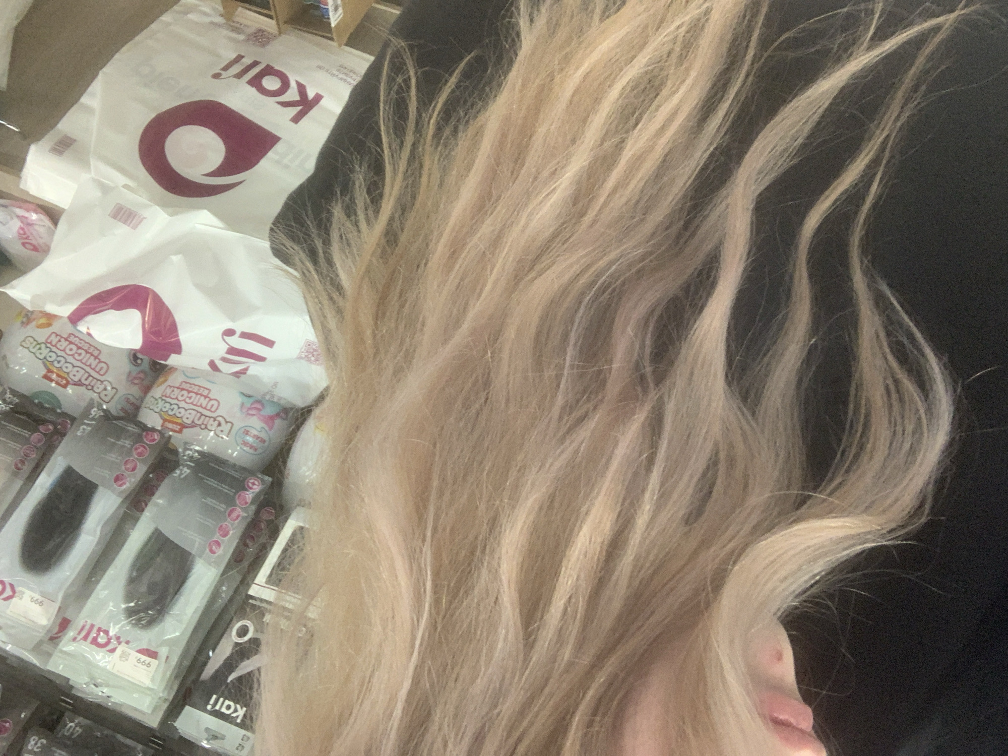 The image features a close-up view of a person's long, wavy blonde hair in the foreground, partially obscuring the scene behind it. The person's chin and mouth are visible in the bottom right corner.
The background, likely a store interior in Saint Petersburg, Russia, shows various retail items. Prominently displayed are several white shopping bags with a dark pink/maroon "kari" logo and text. Some bags have barcodes. Below these, there are packaged goods, including egg-shaped containers labeled "RainBocorns UNICORN RESCUE." Other packaged items, possibly hair extensions or small accessories, are visible in clear plastic. These packages feature the "kari" logo and various numbers such as "38", "40", "42", "43", "45", which likely indicate sizes. One package also shows "999.", possibly a price.
The activity appears to be the person browsing or being near these products, possibly during a shopping trip. The lighting suggests an indoor environment. Visible text includes the brand "kari," "RainBocorns UNICORN RESCUE," and various numbers and a price. Partially visible Russian text such as "РУЗИ" and "АНИРУЙТЕ О" can be discerned on some of the shopping bags.