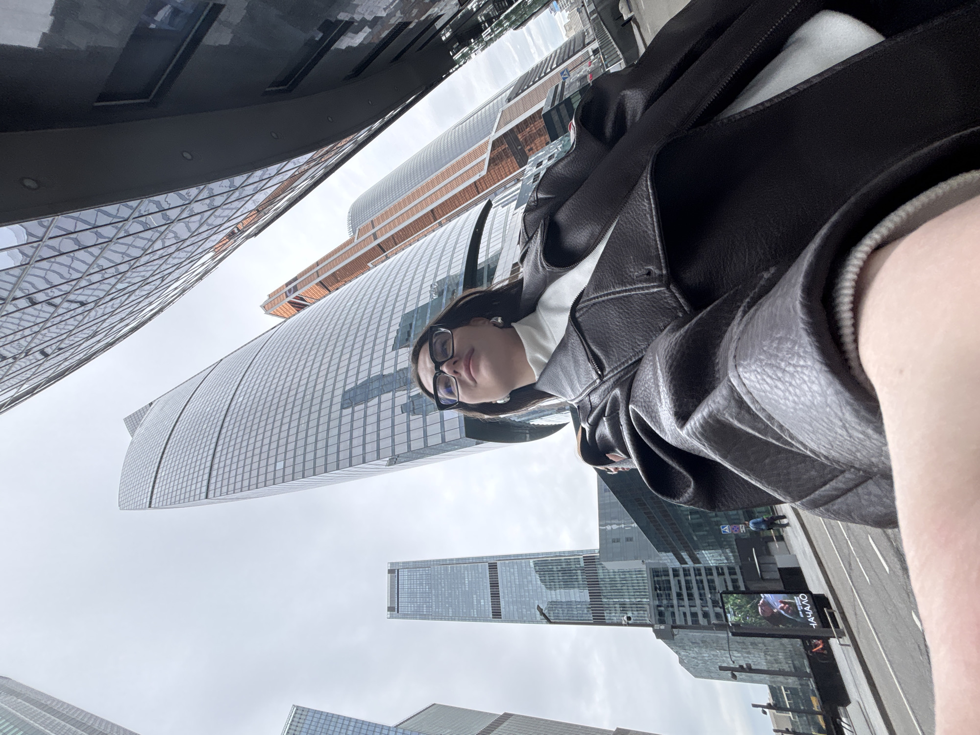 The image shows a view of the Moscow International Business Center, commonly known as "Moscow City," in Moscow, Russia. The shot appears to be taken by someone looking upwards amidst the skyscrapers. The woman is wearing eyeglasses and is partially visible in the frame, and is wearing a dark leather jacket. She's looking at the camera. Skyscrapers are seen from a low angle. A billboard in Russian is visible. The sky is overcast.