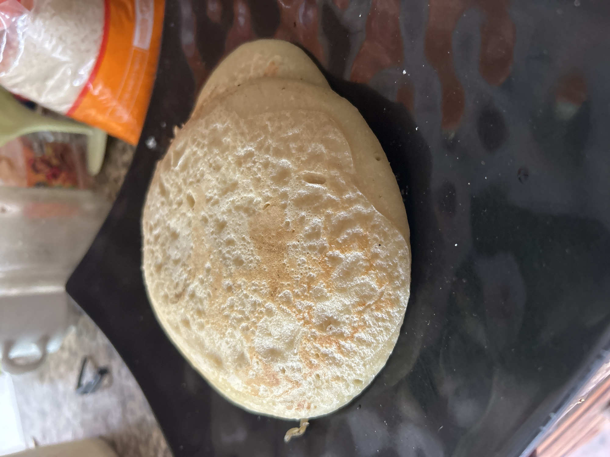 The image shows two light brown pancakes stacked on a black plate. The pancakes have a textured surface with small bubbles and some slightly browned areas. The plate is placed on a countertop. In the background, out of focus, are a white mug, some kitchen appliances, and a bag of what appears to be rice. The scene is indoors, likely in a kitchen setting. The location of Santa Ana, Venezuela is not depicted in the image.