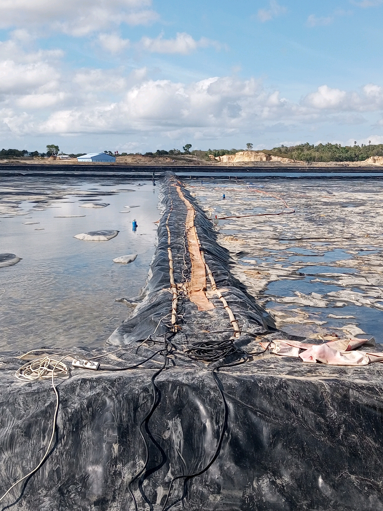 This image depicts an outdoor industrial scene under a partly cloudy sky. In the foreground, a large, dark, impermeable liner covers the ground, with some sections appearing wrinkled and uneven. Several black electrical cables and a white coiled wire are visible on this liner, along with a white electrical extension cord with three sockets. A pinkish fabric is also present on the liner.
The liner extends into the middle ground, where it forms a boundary between a shallow body of murky water and a more solid, partially dried-up area with a patchy, uneven surface. Several light-colored, irregular shapes, possibly floating objects or debris, are dispersed in the water. A blue object, possibly a cap or marker, is standing upright in the water.
Further in the background, a long, raised embankment is covered with a black liner, with orange material and piping running along its top. This embankment separates the foreground water from another large expanse of patchy, dried material. Beyond this, the landscape consists of sandy soil, vegetation, and a few trees. In the far distance, a light blue building with a white roof, likely industrial or commercial, is visible. The scene is located in Serubeba, Indonesia.