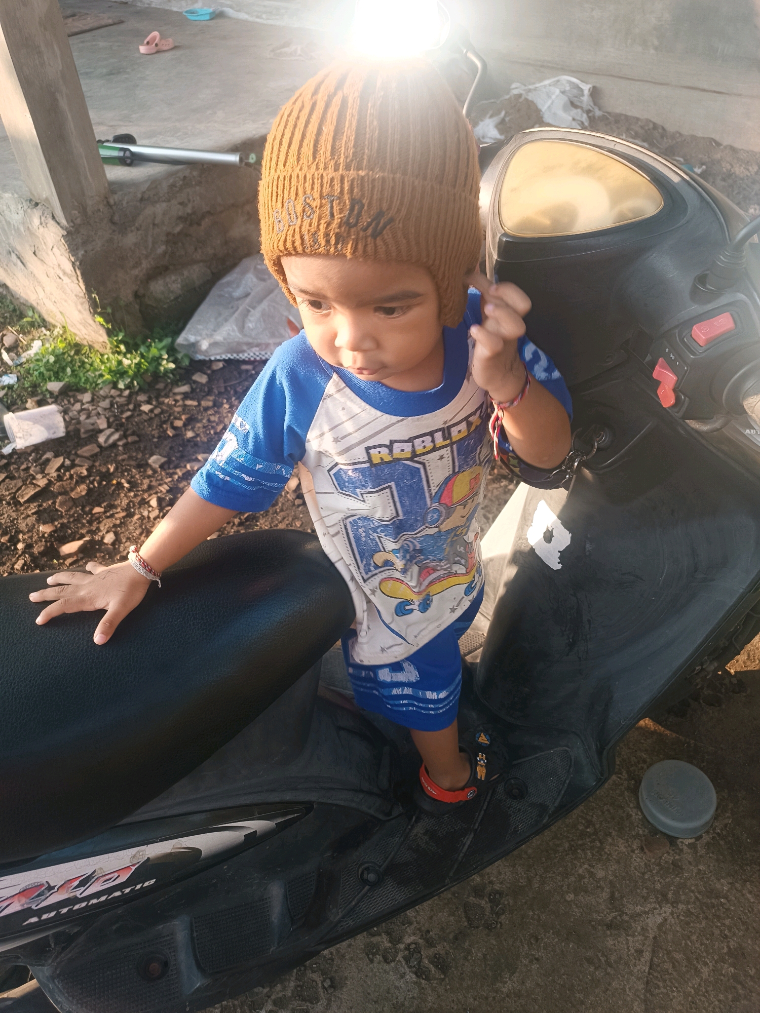 A young child, wearing a brown knitted beanie and a blue and white t-shirt with Roblox graphics, stands on the floorboard of a black scooter. The child's left hand rests on the black seat of the scooter, and their right hand is raised to their head. The child is wearing red and black sandals. The scooter is parked on a paved surface with scattered rocks and dirt. In the background, there is a concrete structure, some greenery, and what appears to be a plastic bag. The image is taken outdoors, likely in a residential area, with the sun casting a bright glare. The location is identified as Tabanan, Indonesia.