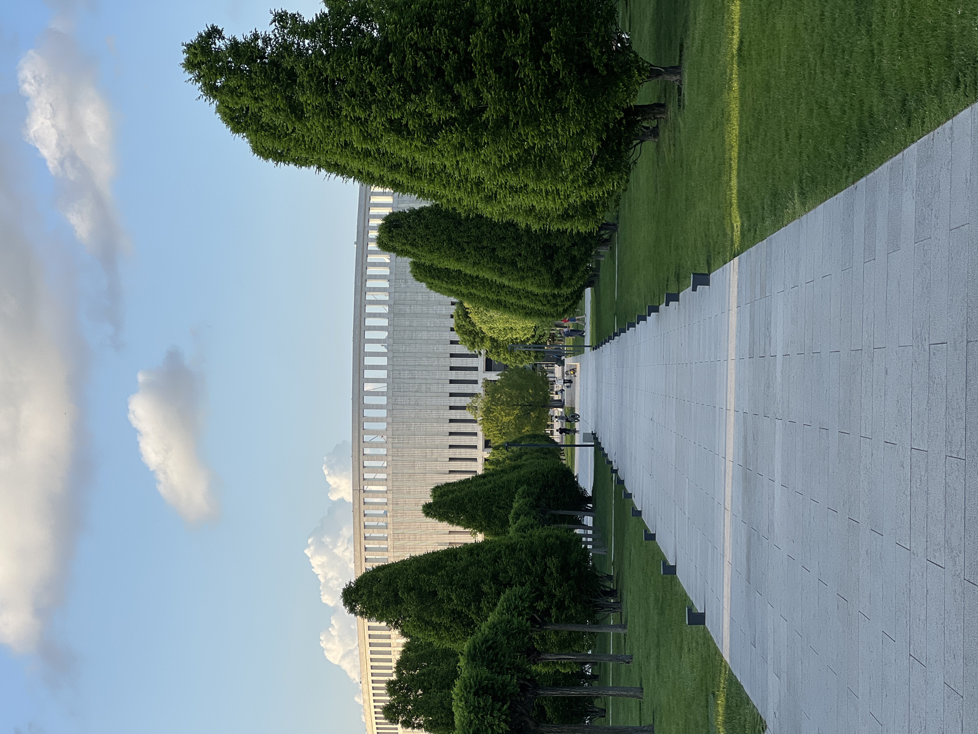 The image shows a perspective view down a wide, grey-tiled walkway lined with evenly spaced, cone-shaped trees. To the left of the walkway, there's a large, modern building with repeating vertical and horizontal elements under a blue sky with cumulus clouds. To the right of the walkway, there's a stretch of green grass. The trees lining the walkway are well-manicured. The scene suggests an urban park environment in Krasnodar, Russia, based on visual similarity to images of Krasnodar Park. Some people are faintly visible further down the walkway.