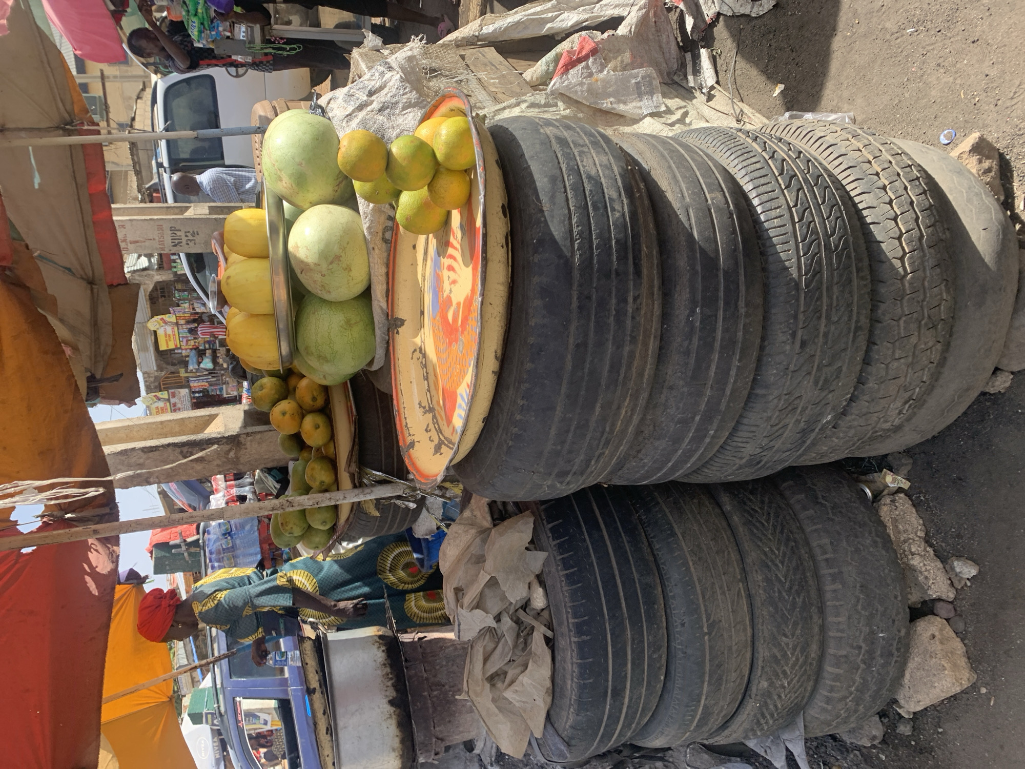 The image shows a street scene in Rafin Par, Nigeria. In the foreground, a stack of used tires is visible. Adjacent to the tires is a display of produce including oranges, yellow melons, and watermelons. The produce is arranged on a stand with a metal tray featuring a colorful design. Further back, a woman wearing an orange headscarf and patterned clothing is visible at her stand under a sunshade. In the background, there is a street with people, vehicles, and various shops. The environment appears to be a busy marketplace with a mix of commercial activities.