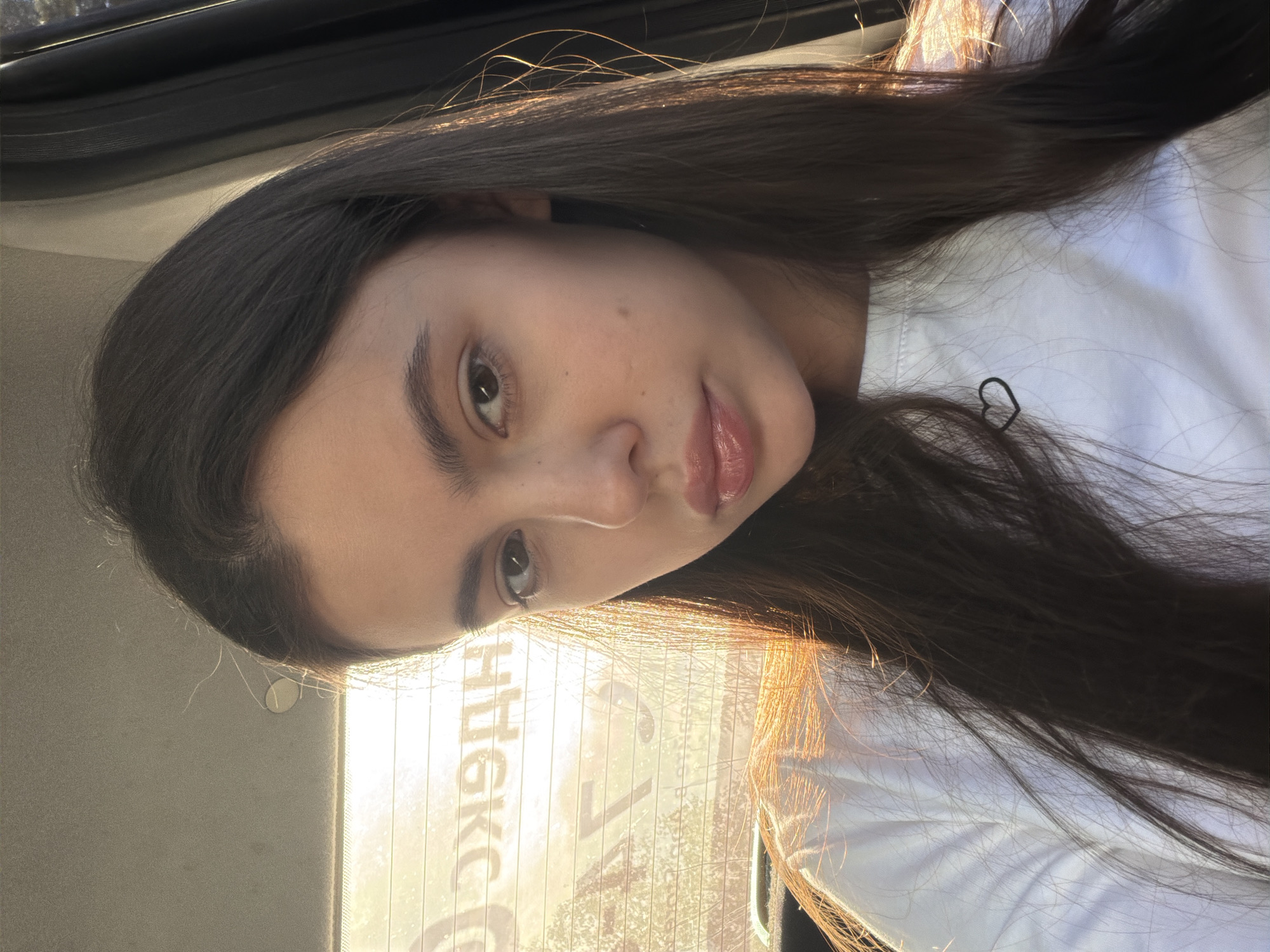 A young woman with long, dark hair is in the backseat of a car. She is wearing a white shirt. The sun is shining through the back window, which has the city name "Чайковский" (Chaikovsky) reflected in reverse on the glass. The location is most likely Chaikovsky, Russia, based on the writing on the window. She has a hair clip shaped like a heart.