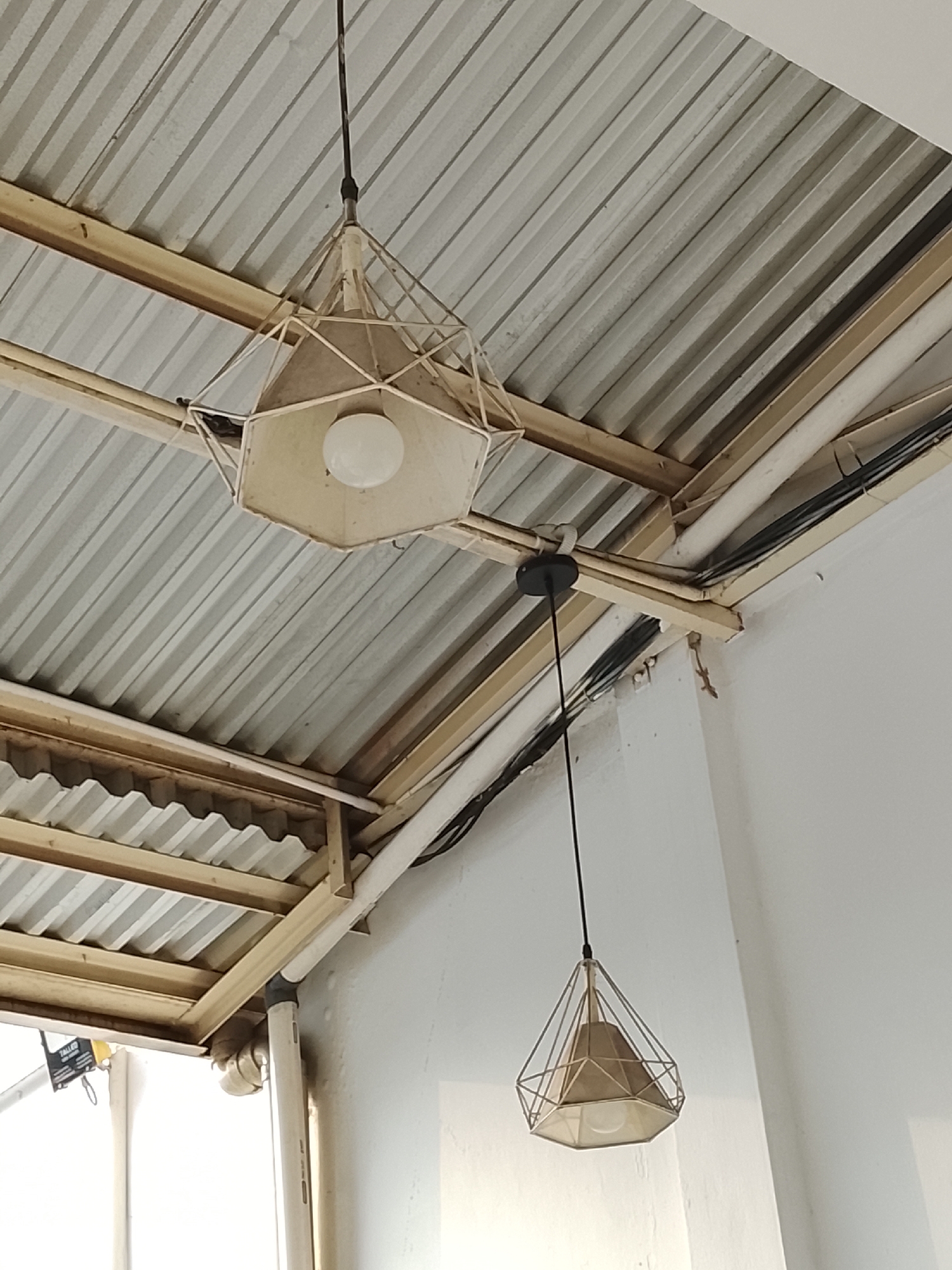 In Kedungwuluh, Indonesia, two geometric pendant lamps hang from a corrugated metal ceiling supported by wooden beams. The larger lamp, closer to the camera, features a hexagonal shade and an exposed light bulb within a white metal frame. The smaller lamp, further away and suspended by a black cord, has a similar hexagonal shade enclosed in a diamond-shaped metal cage. Wires and pipes are visible along the ceiling structure. A small lizard clings to the white wall to the right of the frame. The overall scene suggests an indoor or semi-outdoor space with a rustic or industrial aesthetic.