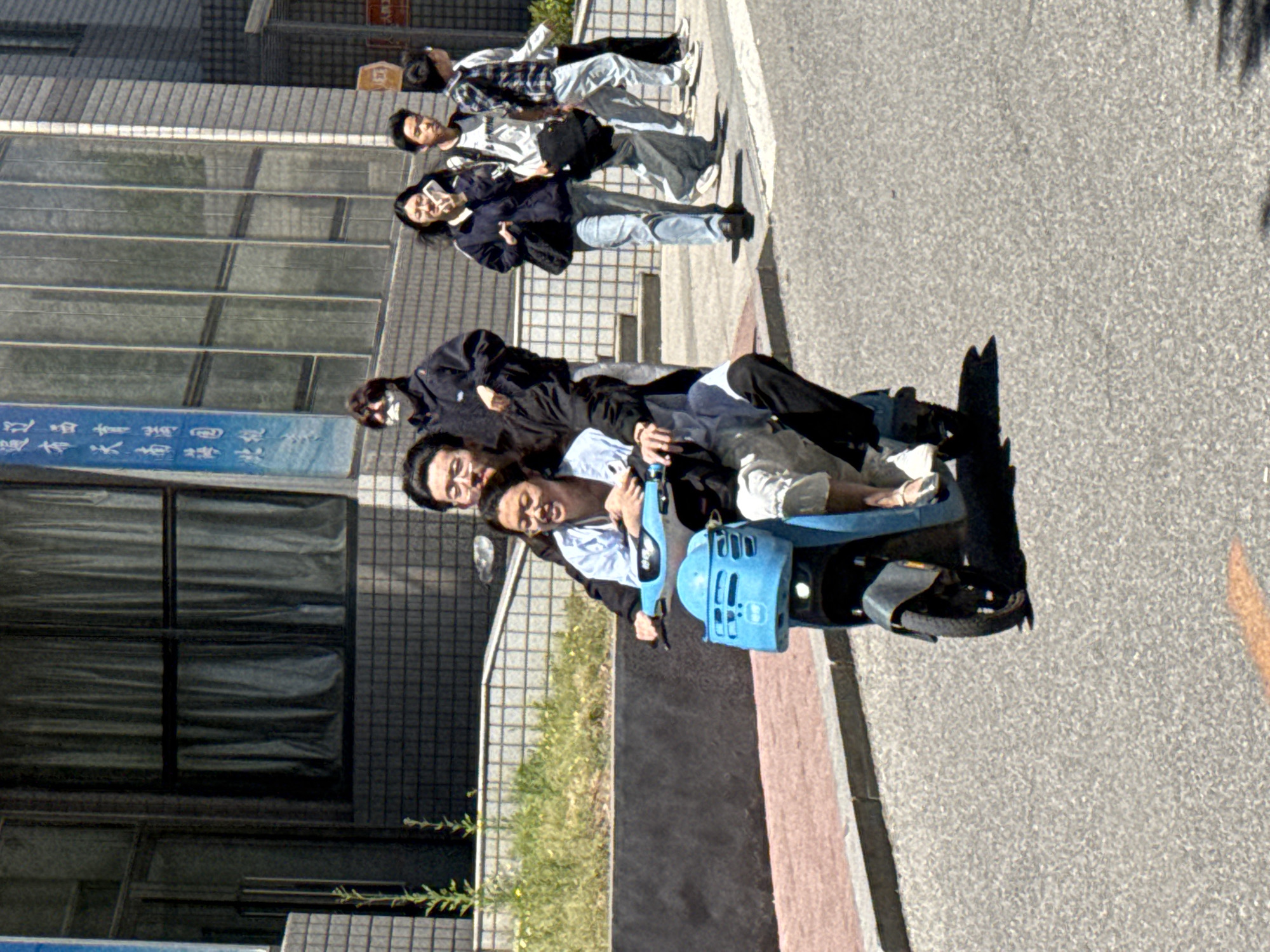 The image shows a group of people. In the background, there are two groups of walking people, and in the foreground, there are four people. They are all crowded together on a single-wheel scooter and sitting on it. The single-wheel scooter is blue. The people are on a paved road in front of a building. The sign above the building reads "Zhongtun, China".