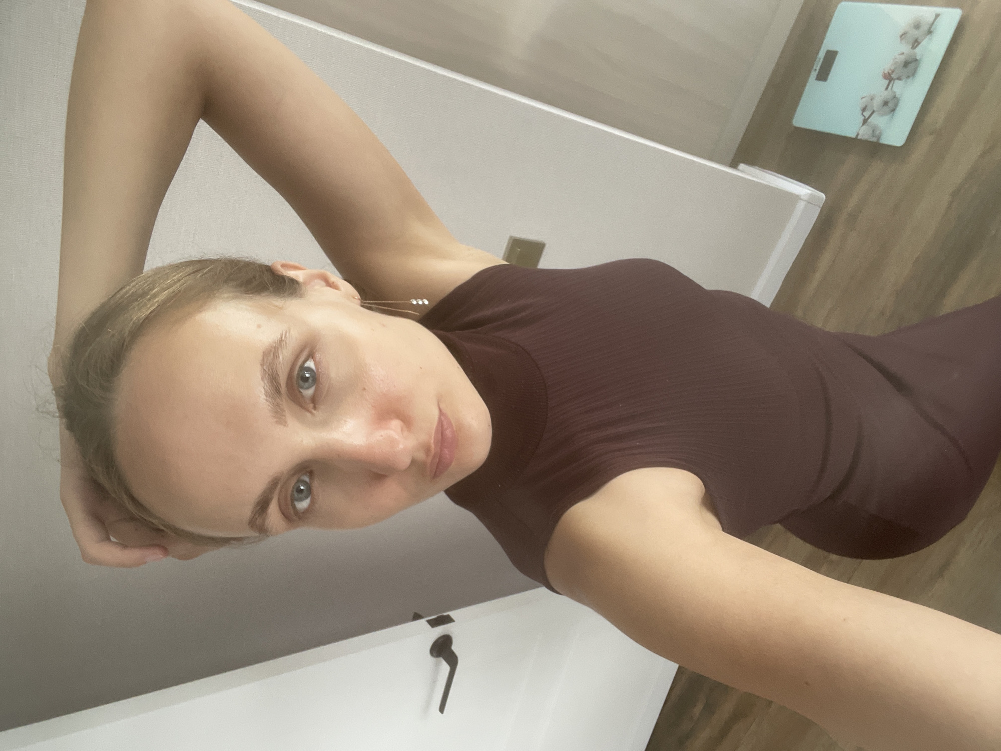 This is a slightly angled, full-body selfie of a young Caucasian woman lying on her side on a wooden floor. She is wearing a dark plum-colored, ribbed, form-fitting dress. Her left arm is bent and raised, with her hand resting on the back of her head. Her right arm is bent with her hand on her waist. She has light brown hair, light blue eyes, and fair skin. Her gaze is directed at the camera with a neutral expression. A digital scale with a cotton branch design is visible on the floor behind her to the right. To her left, a white door with a black handle is visible. The lighting suggests it is daytime. The location is identified as Podshivalovo, Russia.