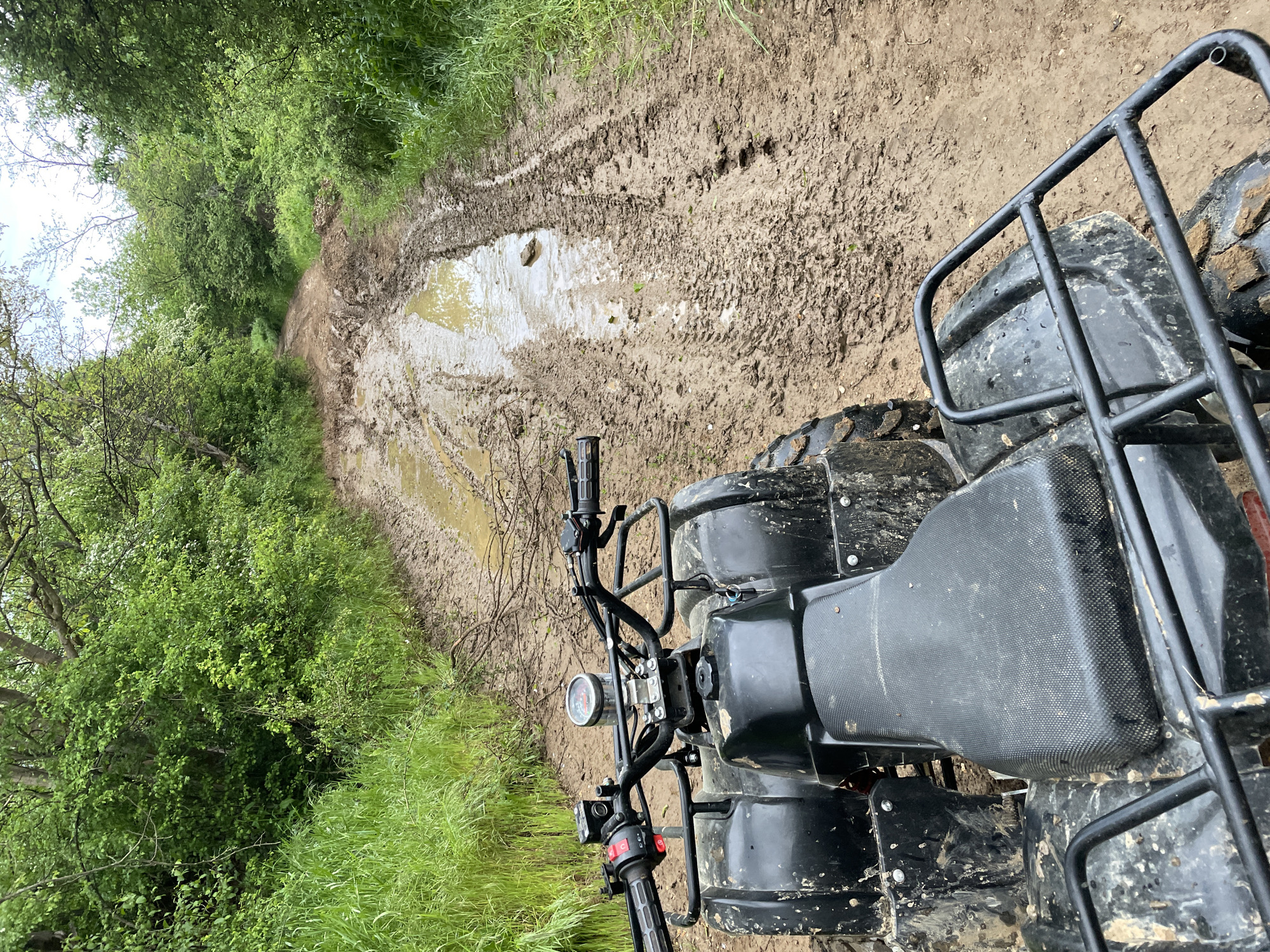 A black ATV is positioned on a muddy trail, facing forward. The trail is deeply rutted with water-filled depressions. Green vegetation lines both sides of the trail. The location is Unknown, Unknown.