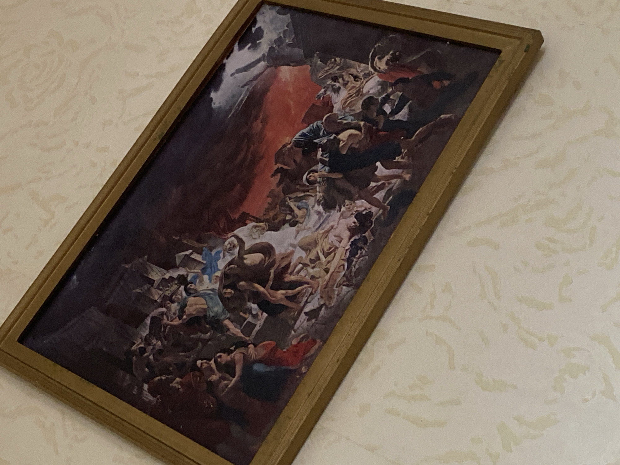 The image depicts a framed painting hanging on a wall with a patterned, off-white surface. The painting, which is rectangular and tilted diagonally, features a chaotic scene with numerous figures rendered in a classical style. The figures are predominantly light-skinned and appear to be in various states of distress, with some intertwined and others falling or ascending. The color palette is dominated by dark reds, browns, and whites, creating a dramatic and turbulent atmosphere. The figures appear to be in some sort of tumultuous event, with a darker mass above and a lighter mass below. The frame around the painting is gold-colored.