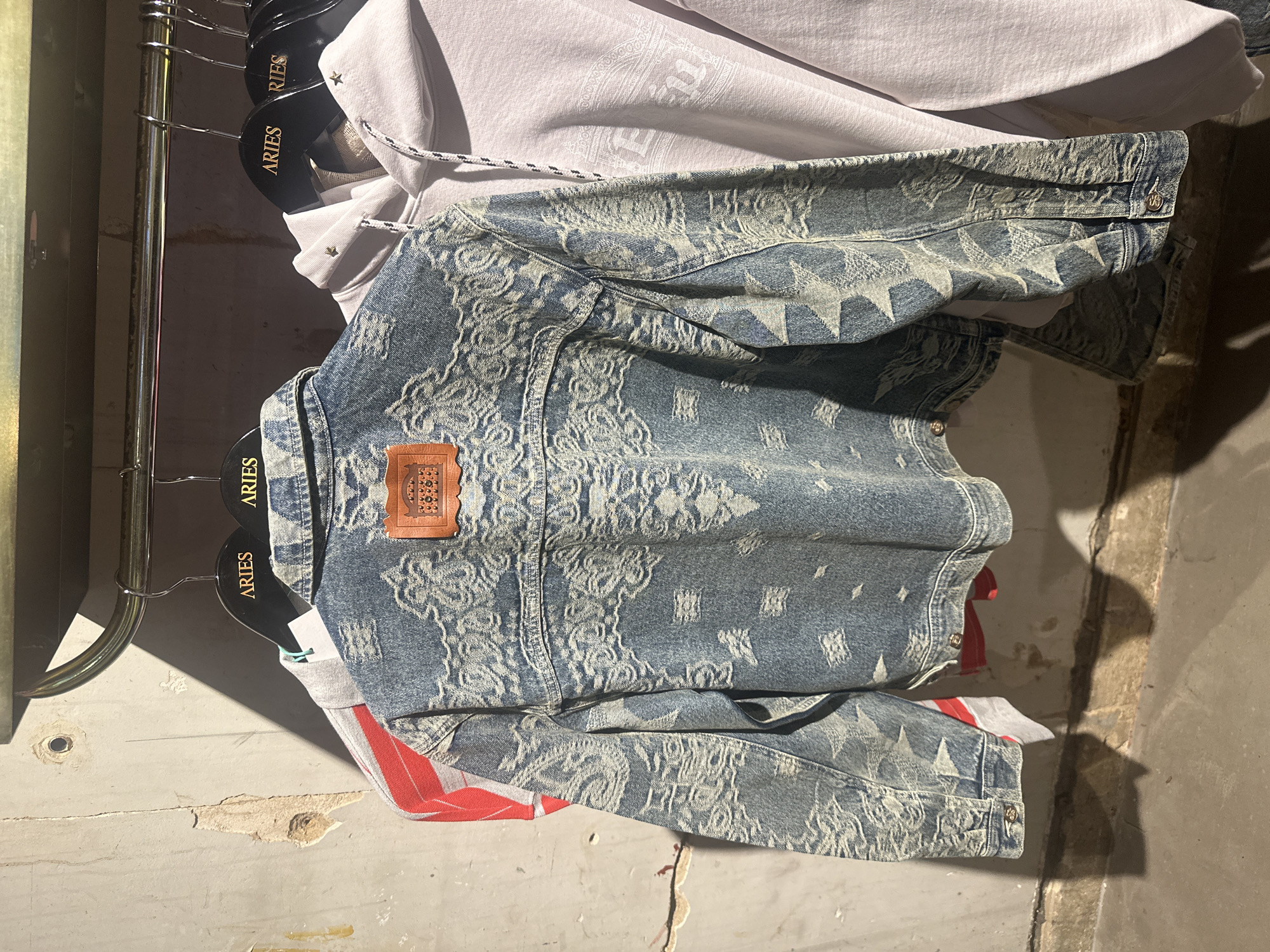 This media file captures an indoor scene, likely within a retail store or a display area, featuring several garments hanging on a rack. The primary subject is a blue denim jacket with an intricate, light-colored pattern that includes stars and ornate designs, giving it a distressed or vintage aesthetic. A small brown leather patch with an embossed design is visible on the upper back of the jacket.
Behind the denim jacket, a light-colored (possibly white or light grey) hoodie is hanging, partially visible. This hoodie also features subtle patterns and small star-shaped embellishments. Its drawstring is visible, and the fabric appears soft. Below the denim jacket, hints of another garment in red and white stripes can be seen. All these items are suspended on black hangers, each bearing the visible text "ARIES" in gold lettering. The hangers are placed on a metallic clothing rack.
The background consists of a rustic or industrial-looking wall, painted a light, neutral color, possibly off-white or beige. Parts of the wall show signs of wear and tear, with chipped plaster and exposed rougher textures. A small, dark hole or indentation is visible on the wall to the left. The lighting creates distinct shadows, suggesting a strong, focused light source, possibly artificial, illuminating the garments and casting shadows on the wall and floor. The floor appears to be a concrete or similar hard surface, also showing some imperfections. No people are visible, and no specific activity beyond the display of clothing is taking place. The time of day is not explicitly discernible but appears to be well-lit, either by artificial light or natural light from an unseen window.