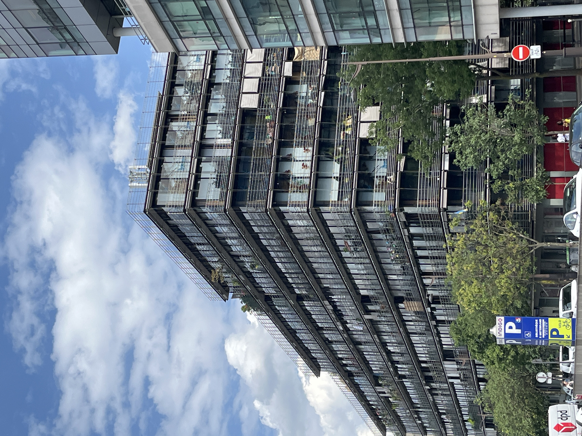 The image shows a modern, multi-story building with a glass facade and horizontal metal accents. The building appears to be an office or residential complex, with visible balconies or terraces on some floors, some of which are adorned with plants. Adjacent to the building, there are several trees, some of which are quite large and leafy, suggesting a daytime scene in a temperate climate, likely during the spring or summer. The sky is blue with scattered white clouds, indicating fair weather.
In the foreground and to the right, a white van with "dpd" logo is partially visible. There's also a blue and yellow sign for "indigo" with a "P" symbol and accessibility icon, indicating a parking area. Another sign above it has a red circle with a diagonal line across it and a bicycle symbol, suggesting a "no cycling" or similar restriction. There is also a white van with the word "velo" on it. The scene suggests an urban environment with commercial or residential activity. There are some people visible in the reflection of the building or in the background, but they are not a primary focus and are too indistinct to identify specific activities. The overall impression is one of a well-maintained, contemporary urban landscape.