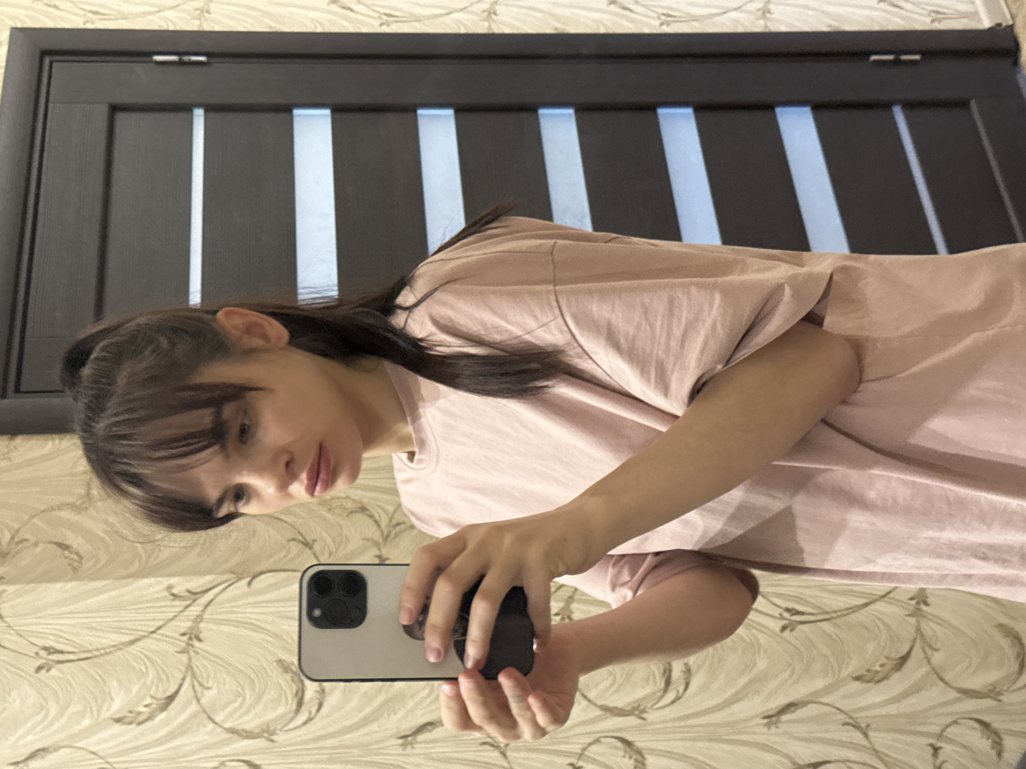 A young woman is taking a selfie in what appears to be an interior setting. She is holding a smartphone with a triple-lens camera, likely an iPhone, in her left hand. She has dark hair with bangs and is wearing a light pink t-shirt. The background features a patterned wallpaper and a dark door with light panels. There is no information in the image to confirm that the photo was taken in Rostov-on-Don, Russia. The location is unknown.