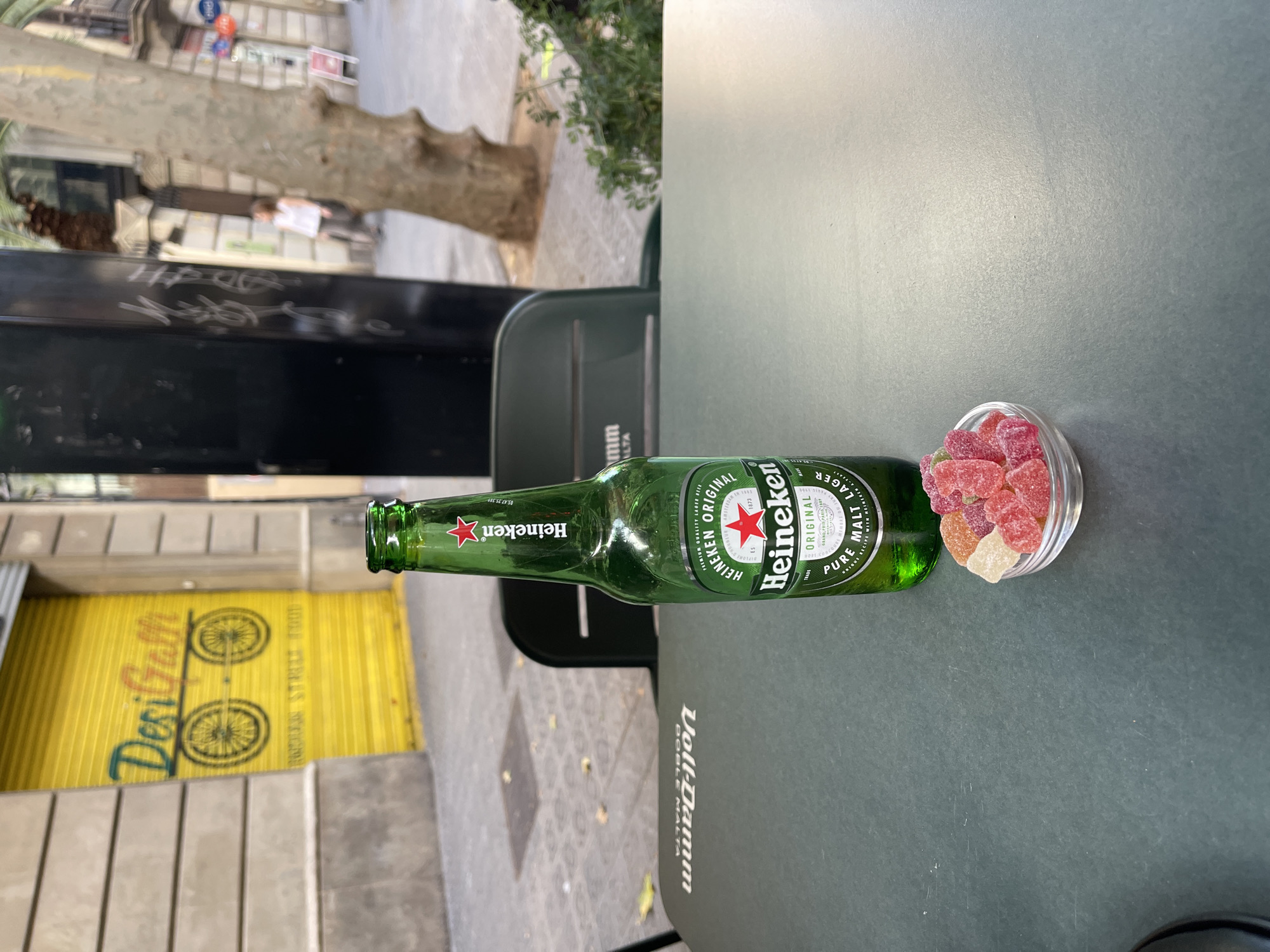 A green glass bottle of Heineken beer and a small glass bowl filled with colorful gummy candies are placed on a dark, textured table outdoors, with a backdrop of a city street. The Heineken bottle is upright and partially filled with liquid, its label clearly visible, featuring the brand's iconic red star. The bowl contains a mix of red, pink, and yellow gummy candies.
The table surface has a faint white logo, possibly of a beverage company, with the text "Voll-Damm" and "DOBLE MALTA" discernible. In the background, a closed yellow metal storefront with "DesiGalli" written in colorful script and an illustration of two bicycle wheels is visible. This suggests the location is likely a restaurant or shop.
Further in the background, a tree with rough bark stands to the right, and a sidewalk with paving stones is apparent. A person in a white top and dark pants is walking in the distance on the street. The environment appears to be urban, with buildings and street furniture suggesting a city setting in Barcelona, Spain. The overall scene captures a moment of leisure or a casual break, possibly at an outdoor cafe or seating area.