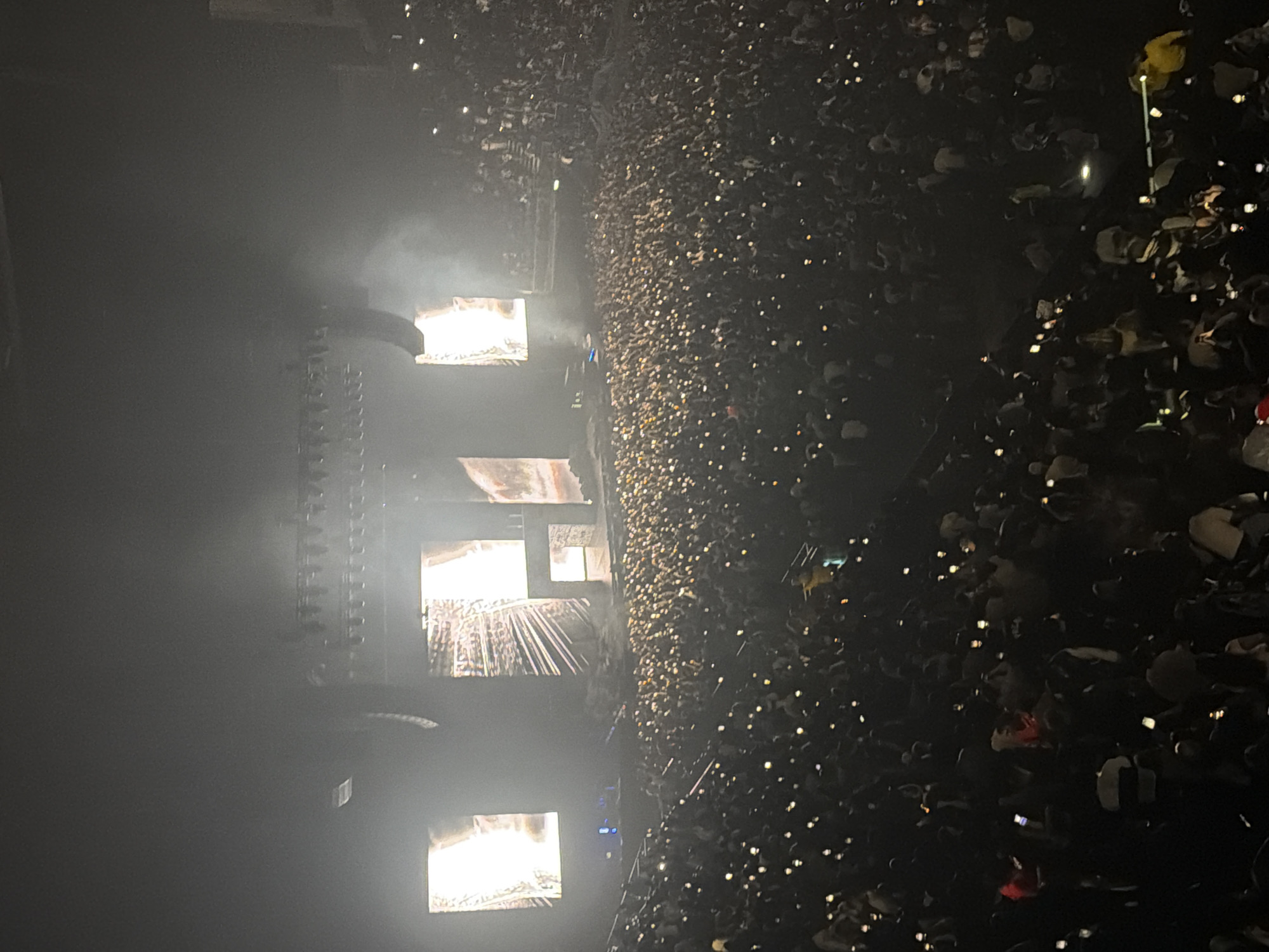 The image depicts a large crowd of people at a concert. Many attendees are holding up their phones, creating numerous points of light across the dark space. Several illuminated screens or stage elements are visible on the left, obscured by smoke or fog. The location appears to be a large indoor arena. While visual details align with large concert venues in London, such as the O2 Arena or Wembley Arena, the image lacks the defining architectural details necessary to confirm a specific London location.