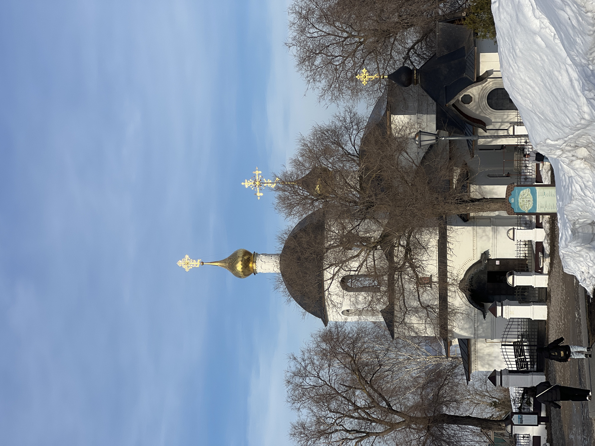 A daylight scene in Sviyazhsk, Russia, depicts the white-walled, multi-domed Dormition Cathedral and Monastery against a clear blue sky. The dominant structure features a dark, hemispherical main dome topped by a white drum, a golden onion dome, and an ornate golden Orthodox cross. Two smaller dark domes, each with a simpler golden Orthodox cross, are visible to the right. The church's facade presents a large, dark, arched entrance beneath a decorative canopy.
In the foreground, two individuals are seen from behind, walking left-to-right on a paved path towards the church entrance. The person on the left wears a dark jacket and pants, while the person on the right wears a dark hooded jacket with a white design on the back and light-colored pants. Bare deciduous trees with visible branches frame and partially obscure the church structure. A white fence with black metal gates and square pillars separates the path from the church grounds. A dark street lamp stands near the fence. To the left of the fence, a blue informational sign with text and a map is displayed. Large mounds of snow are present on the ground, particularly in the lower right quadrant, indicating winter or early spring conditions.