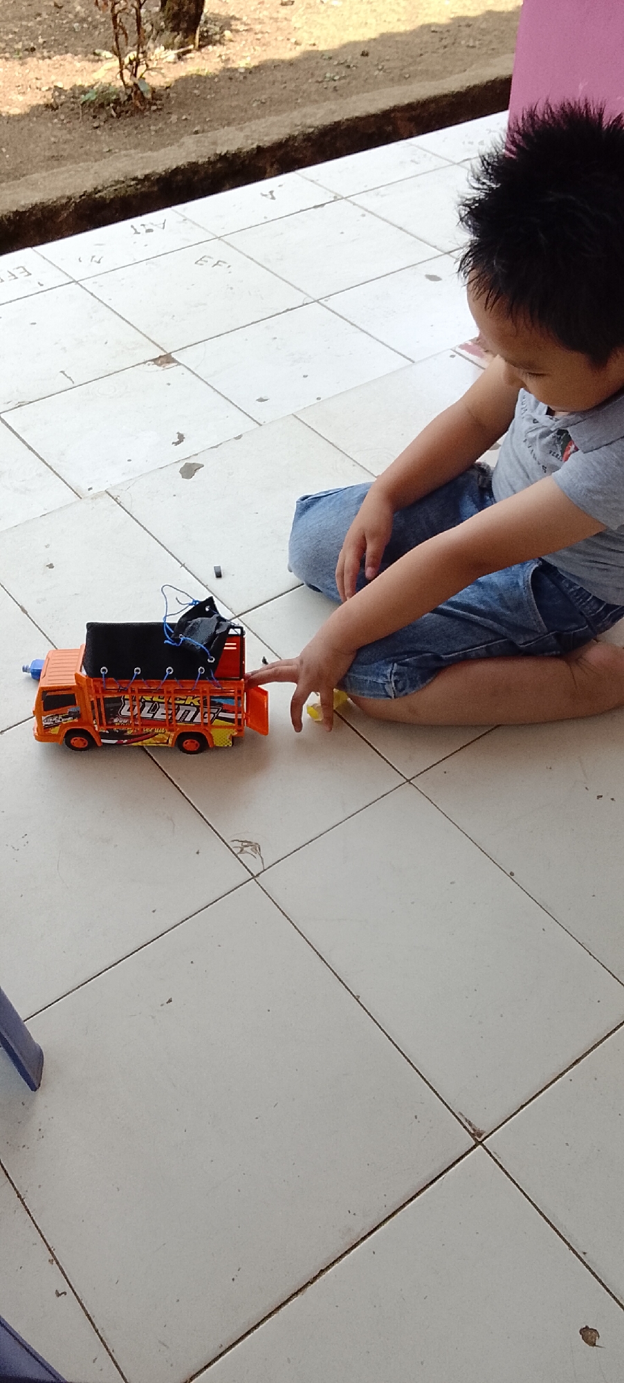 A young child kneels on a tiled floor, playing with an orange toy truck. The truck has a black tarp covering its bed and is adorned with colorful decals. The child is wearing a grey t-shirt and blue denim shorts. The background shows a dirt embankment and some dry vegetation, suggesting an outdoor or semi-outdoor setting. The tiles appear to be standard outdoor patio tiles. The child's right hand is extended towards the toy truck.