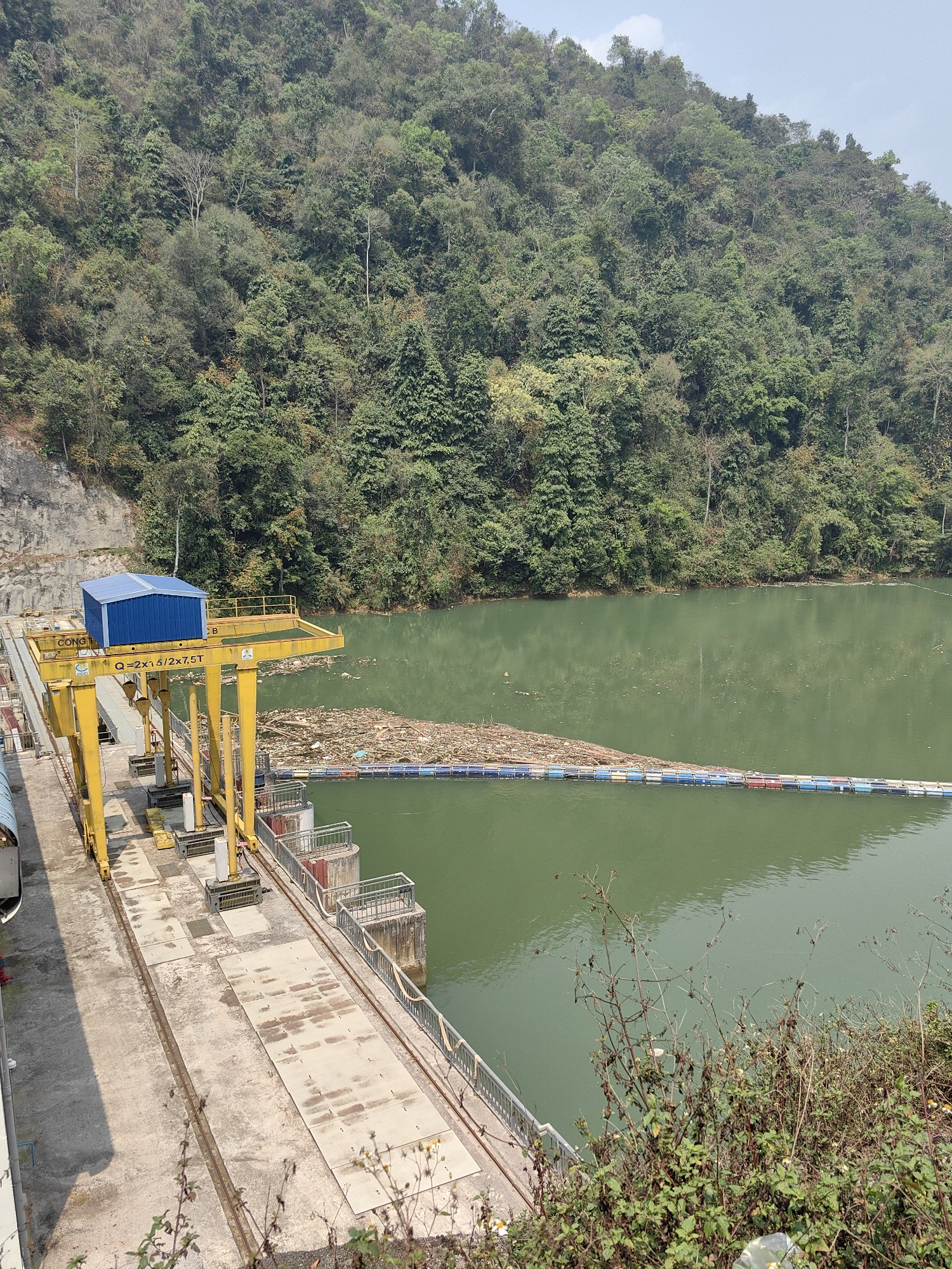 The image depicts a dam in Bảo Lạc, Vietnam. A large body of green water is visible, possibly a reservoir. A barrier made of floating, colorful barrels stretches across the water, collecting debris. A yellow overhead crane structure sits on a concrete surface above the dam's walls. Lush green trees cover the hill behind the dam. Vegetation also grows near the base of the dam in the foreground.