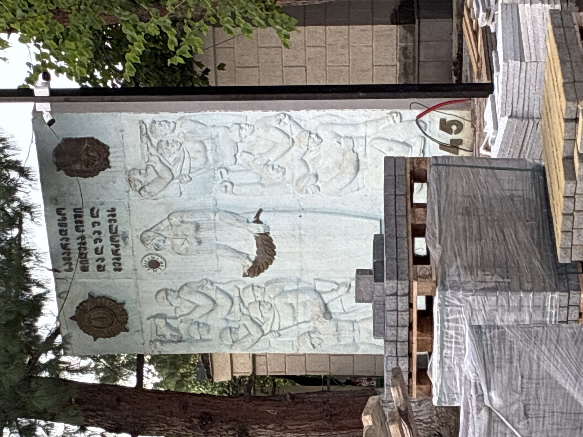 The image shows a weathered, light blue monument with bas-relief sculptures depicting figures, possibly soldiers in battle or laborers. The monument has Georgian script at the top and two circular emblems on either side. In front of the monument, there are stacks of paving stones on wooden pallets and wrapped in plastic, suggesting ongoing construction or renovation work. A dark pole with two security cameras mounted at the top stands to the right of the monument. Behind the monument, a brick wall and trees are visible. A number "45" is painted on the ground near the base of the monument. The scene appears to be outdoors, in what is identified as the city of Surami, Georgia, possibly a public square or park given the monument and surrounding elements.