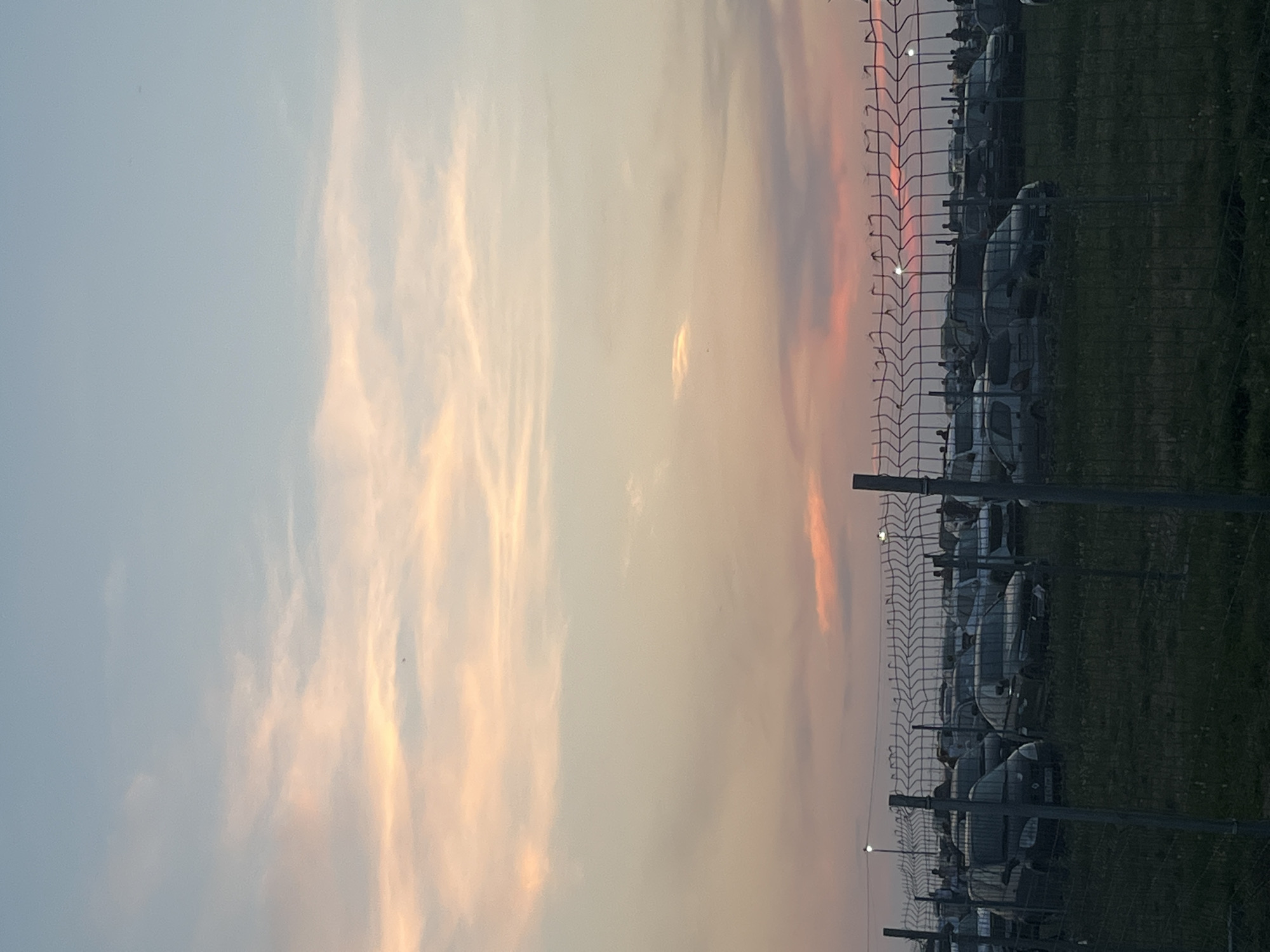 The image depicts an outdoor parking lot filled with numerous cars, observed from behind a wire fence. The sky above is a soft gradient of blue transitioning into hues of pink and orange, indicative of either sunrise or sunset. Several figures, presumably people, are visible amidst the parked vehicles, some appearing to stand or walk around. The ground appears to be covered in grass. The setting is likely the city of Surakovo, Russia, as indicated by the provided context.