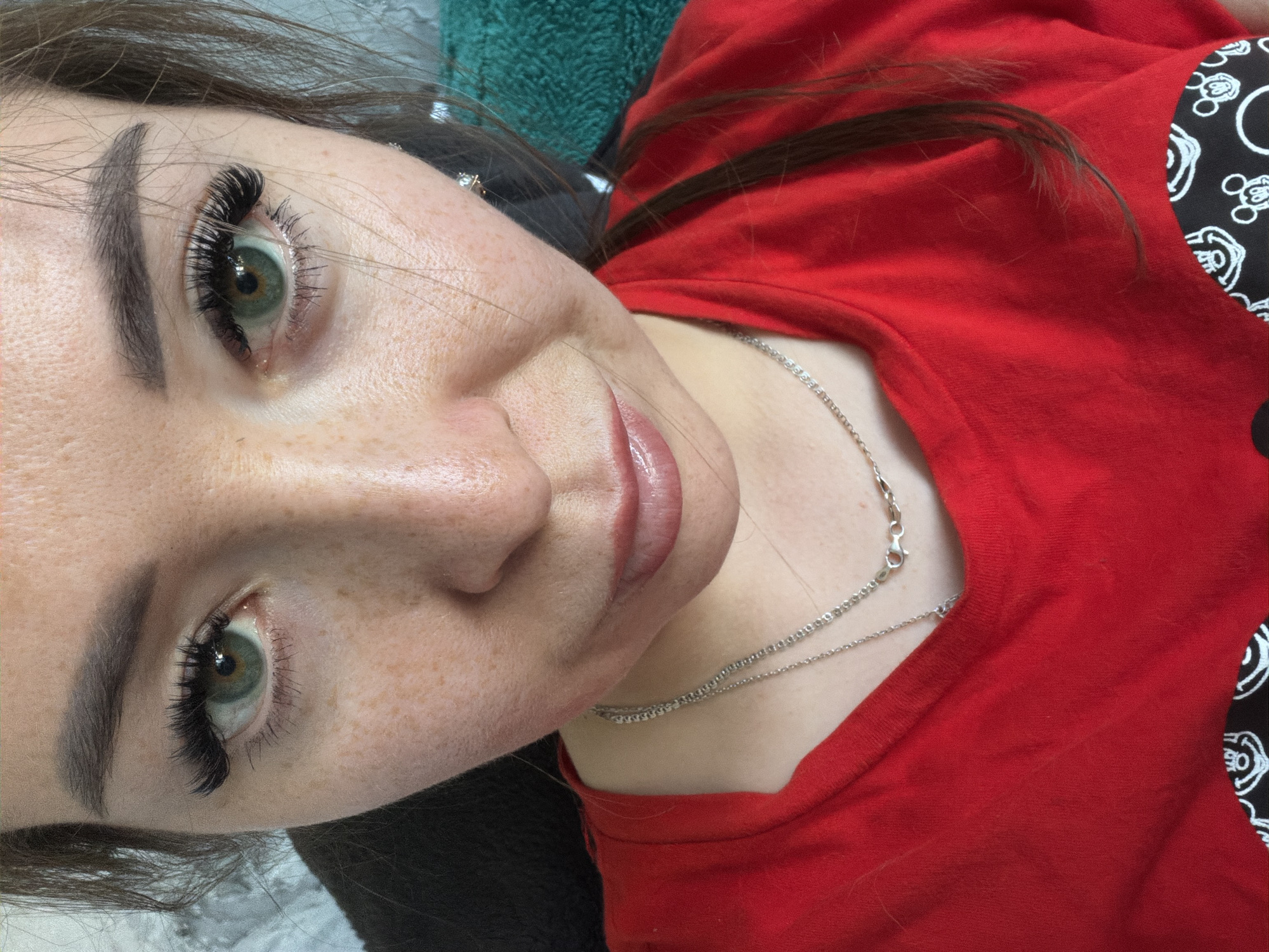 A medium shot shows a Caucasian woman with light skin and freckles, looking at the camera. She has green eyes, dark eyebrows, and dark eyelashes. Her lips are full and appear to be lightly glossed. She is wearing a red t-shirt with two silver necklaces. The background is out of focus, but appears to be an indoor setting with a textured wall on the left and a green textured fabric on the right. This image was taken in Tujmazy, Russia.